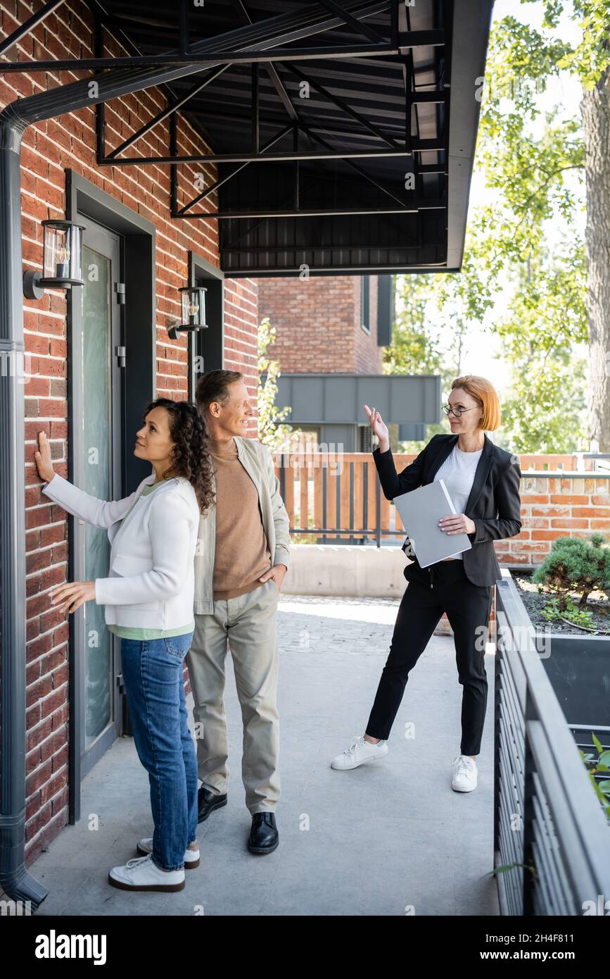 Full Length Of Redhead Realtor In Glasses Pointing At New Modern House