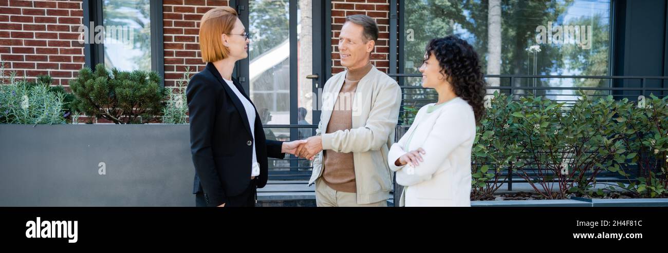 happy realtor in glasses shaking hands with smiling man standing near ...