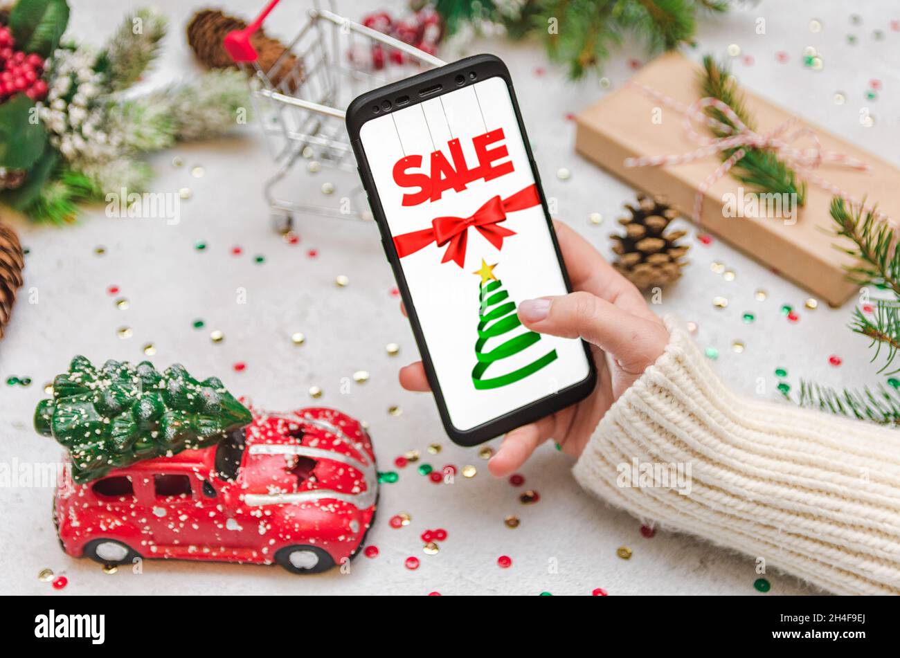Woman in warm and cozy sweater holding a mobile phone and doing some online shopping. Christmas decorations and presents in frame on bright background Stock Photo