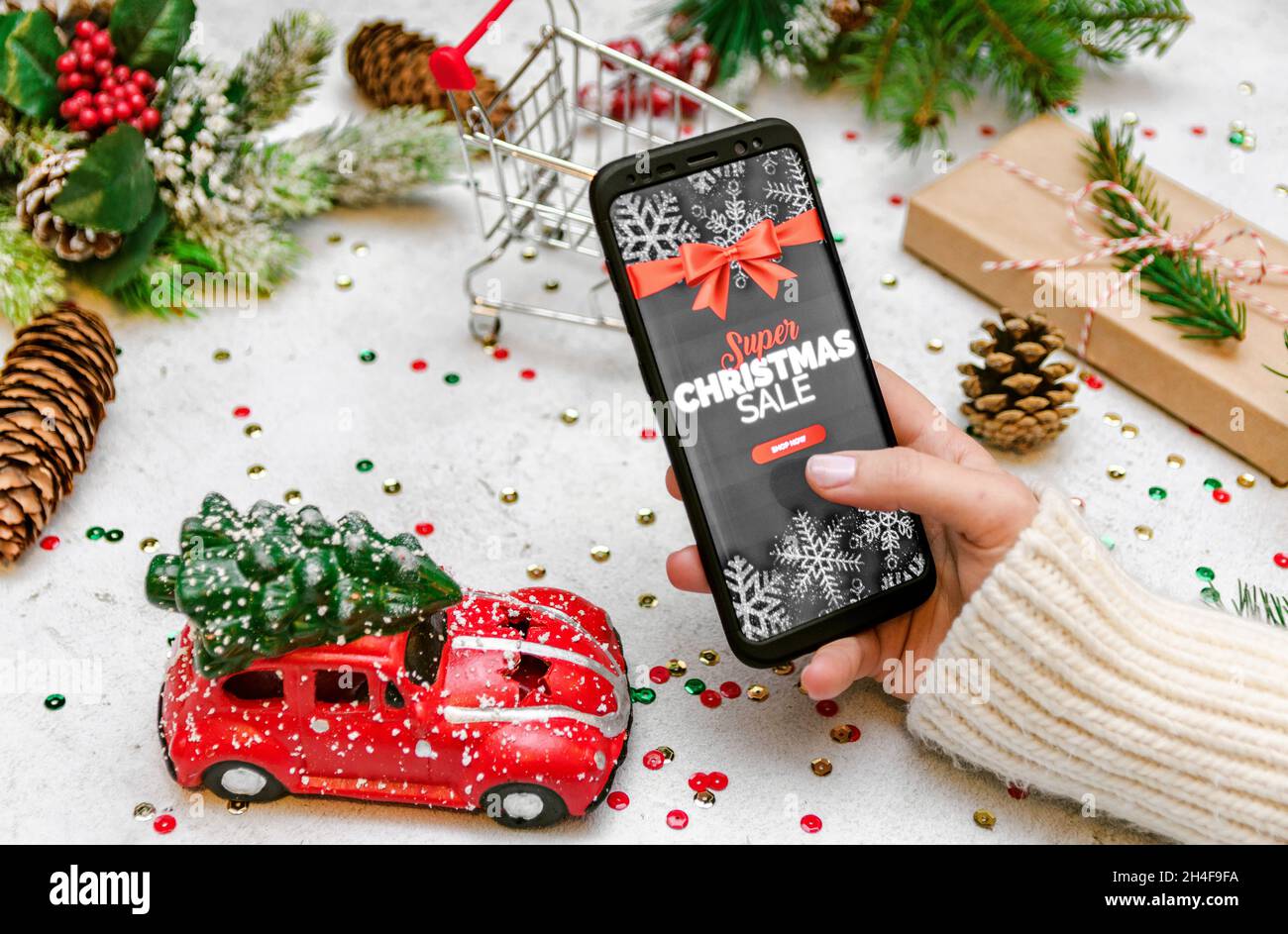 Woman in warm and cozy sweater holding a mobile phone and doing some online shopping. Christmas decorations and presents in frame on bright background Stock Photo