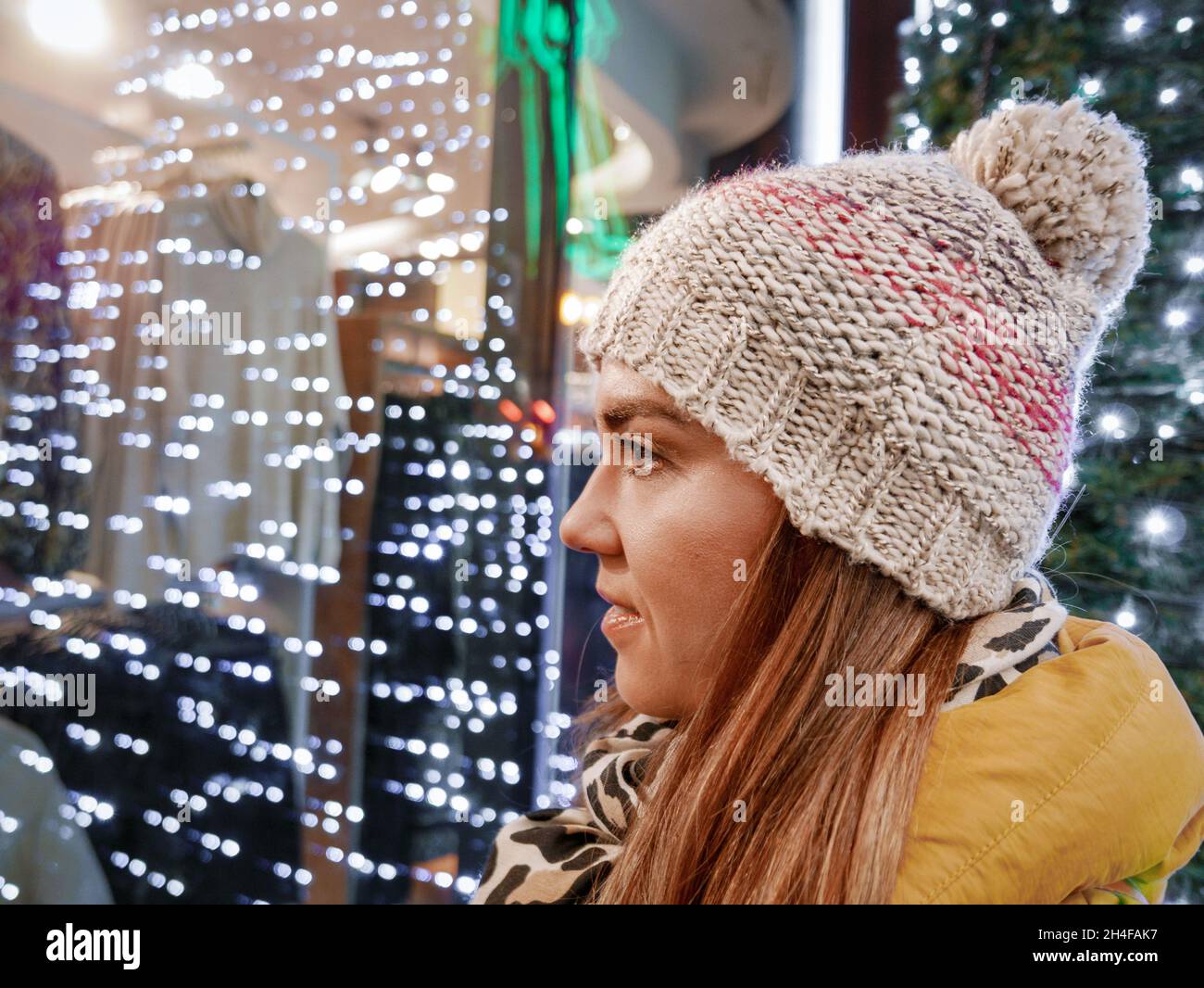 Young woman wearing winter clothes and hat, looking at shop window with christmas lights Stock Photo