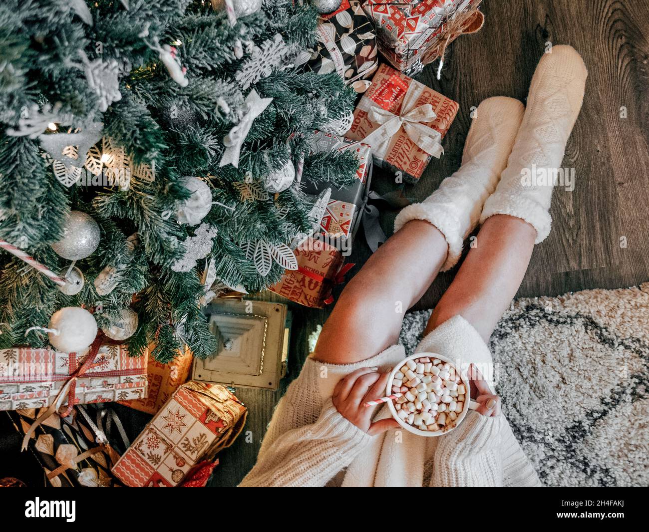 Overhead photo of woman holding hot cocoa with marshmallows, wearing warm socks, sitting by christmas tree Stock Photo