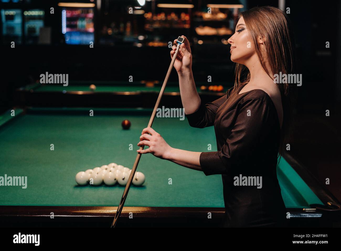 A girl in a dress stands with a cue in her hands and cleans it with chalk in a billiard club with balls in her hands.Playing billiards. Stock Photo