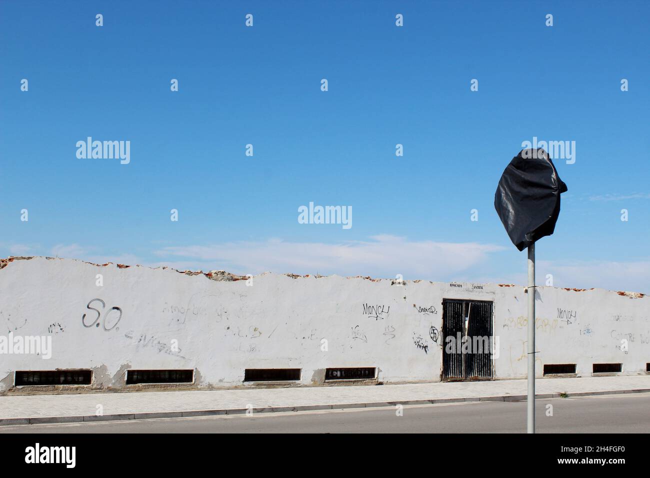 Hell, kaputte Mauer mit Graffitis und einem schwarzen, geschlossenen Tor; davor ein mit  einem schwarzen Müllsack verhülltes Straßenschild, Portugal. Stock Photo