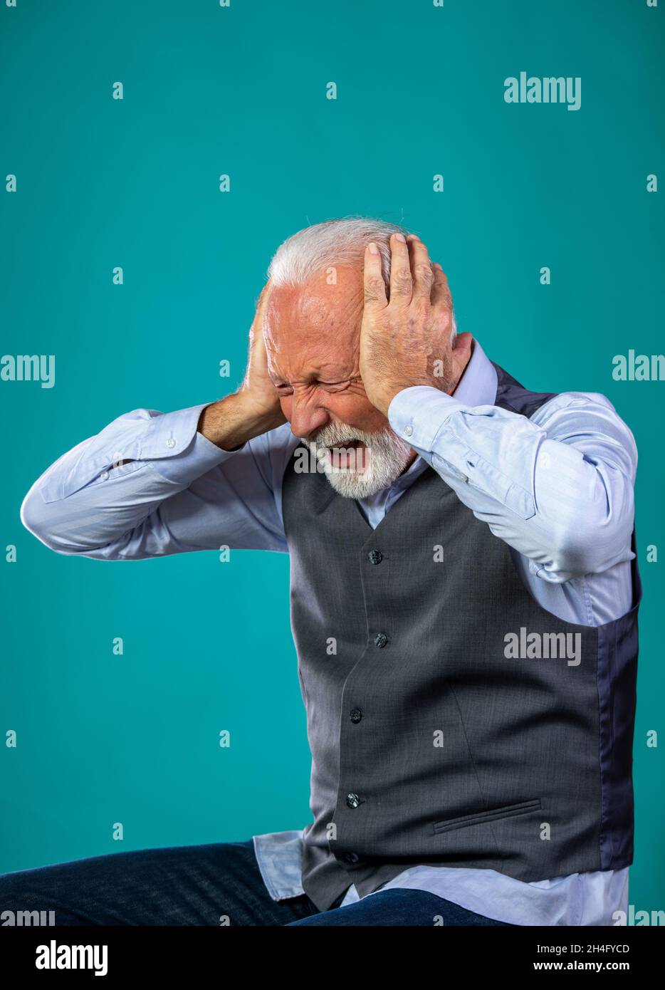 Frustrated old man with white hair and beard holding hands on ears and yelling, on blue background in studio Stock Photo