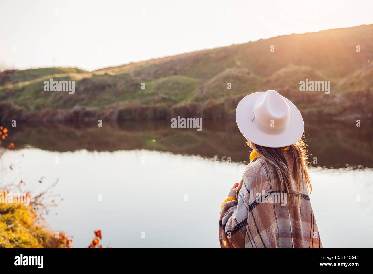 Back view of woman relaxing by autumn river at sunset. Stylish girl in hat enjoying fall landscape in silence. Harmony and balance. Healing nature Stock Photo