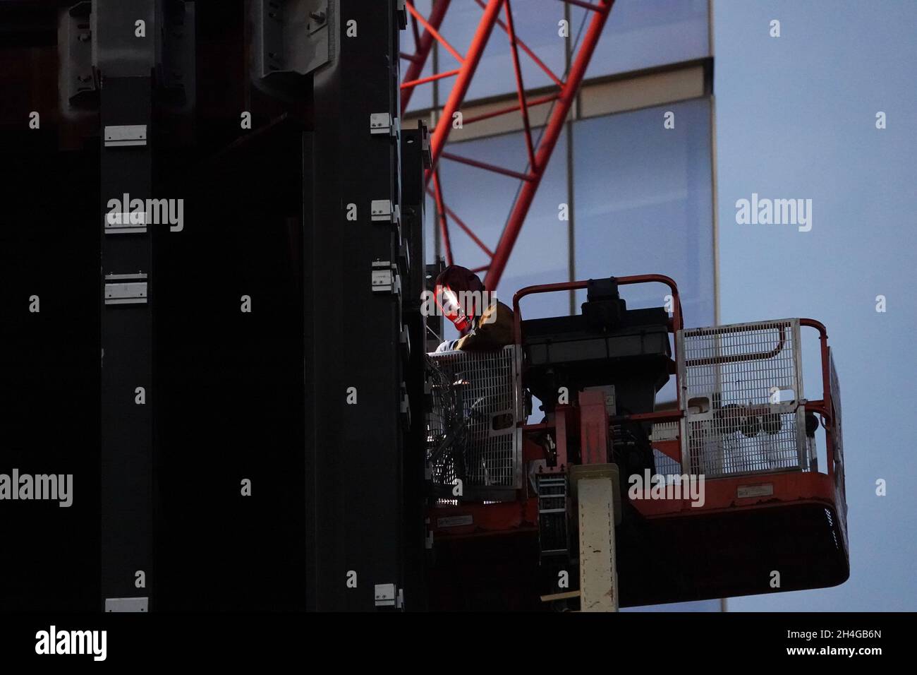 A welders torch lights up the visor of a person welding several stories up on a lift as a new building is constructed Stock Photo