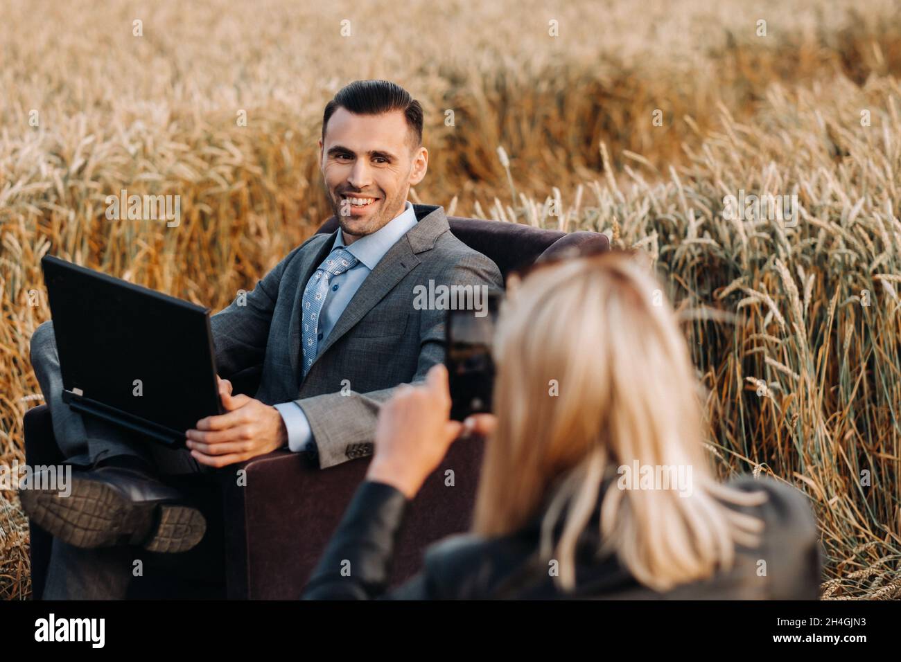 Portrait of a businessman in a suit with a laptop sitting in a chair in a wheat field and photographed by an unrecognizable girl Stock Photo