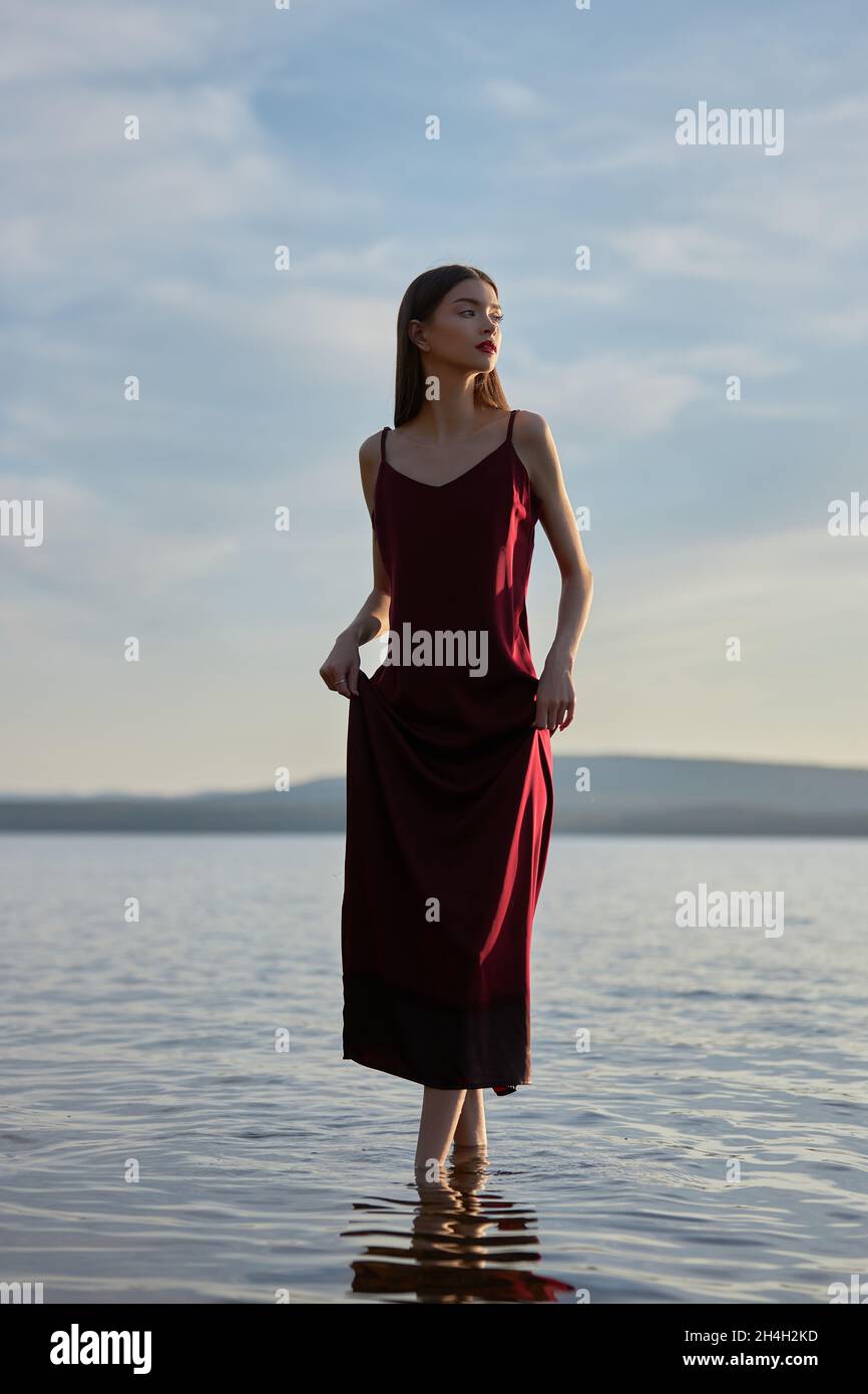 Beautiful woman in light red dress stands in water of lake sea at ...