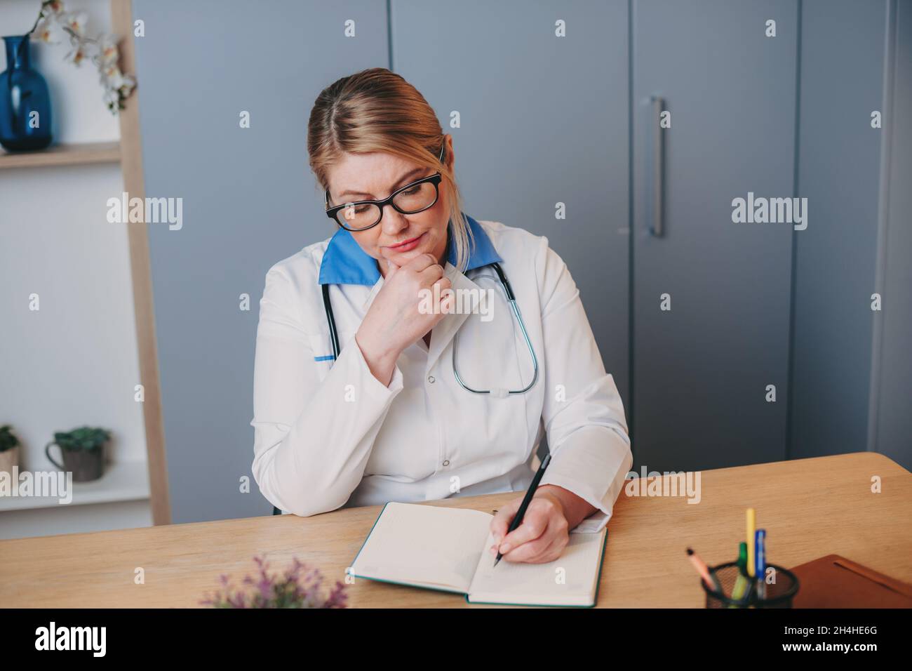 Middle-aged nurse in white clothes is sitting at a desk in hospital and writing something in a notebook. Front view. Stock Photo