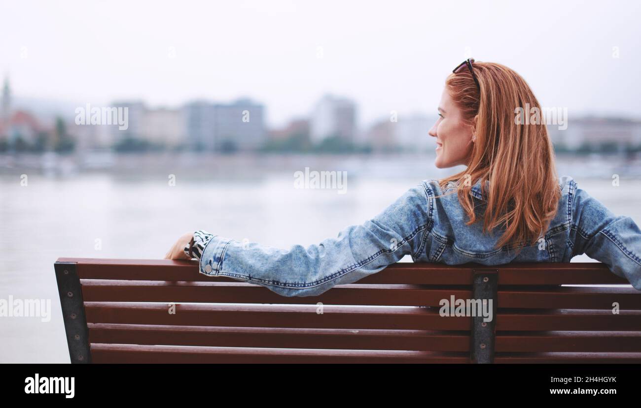 Young redhead woman relaxing on bench at riverside looking side Stock Photo