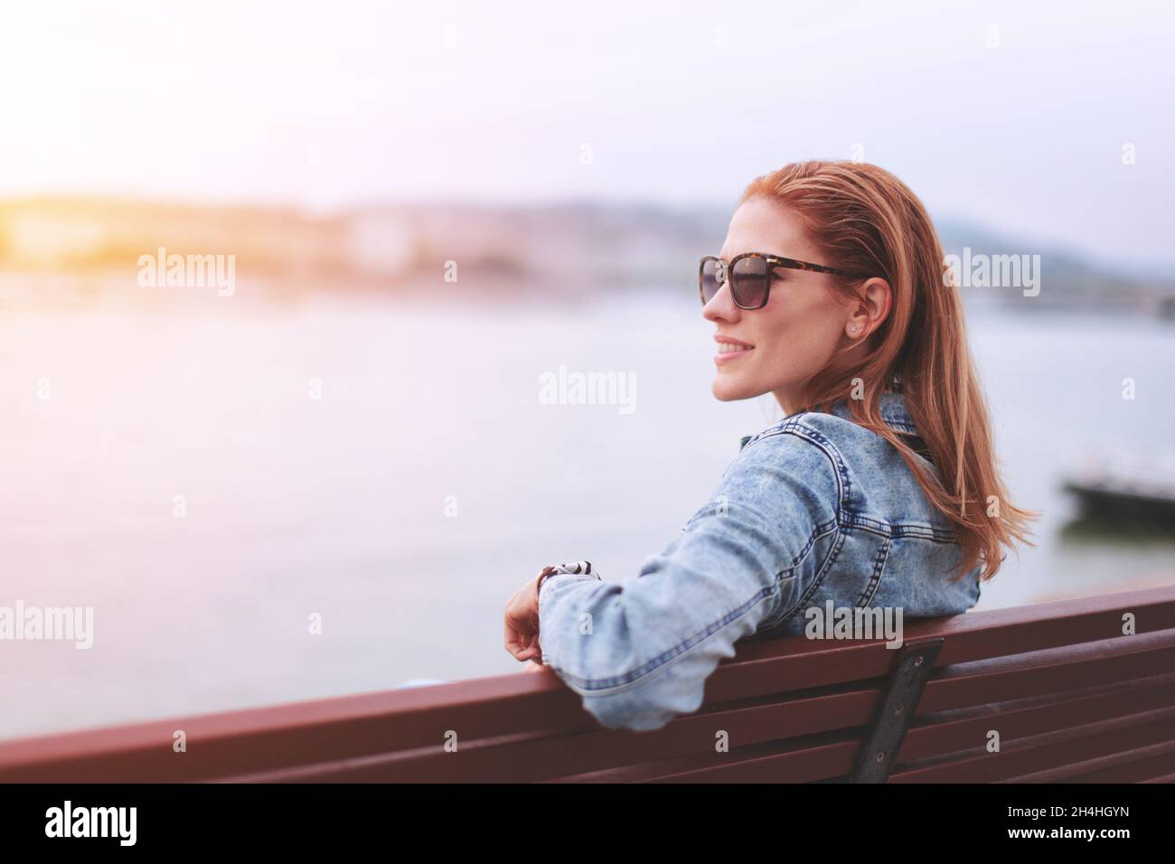 Young balanced redhead Caucasian woman sitting on bench in sunglasses at riverside looking away in sunset Stock Photo