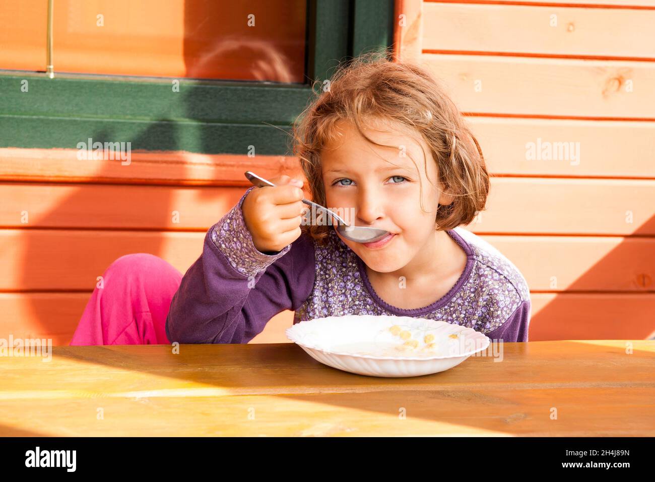 Little girl  eating her milk soup with cereals, sunny outdoors. Stock Photo