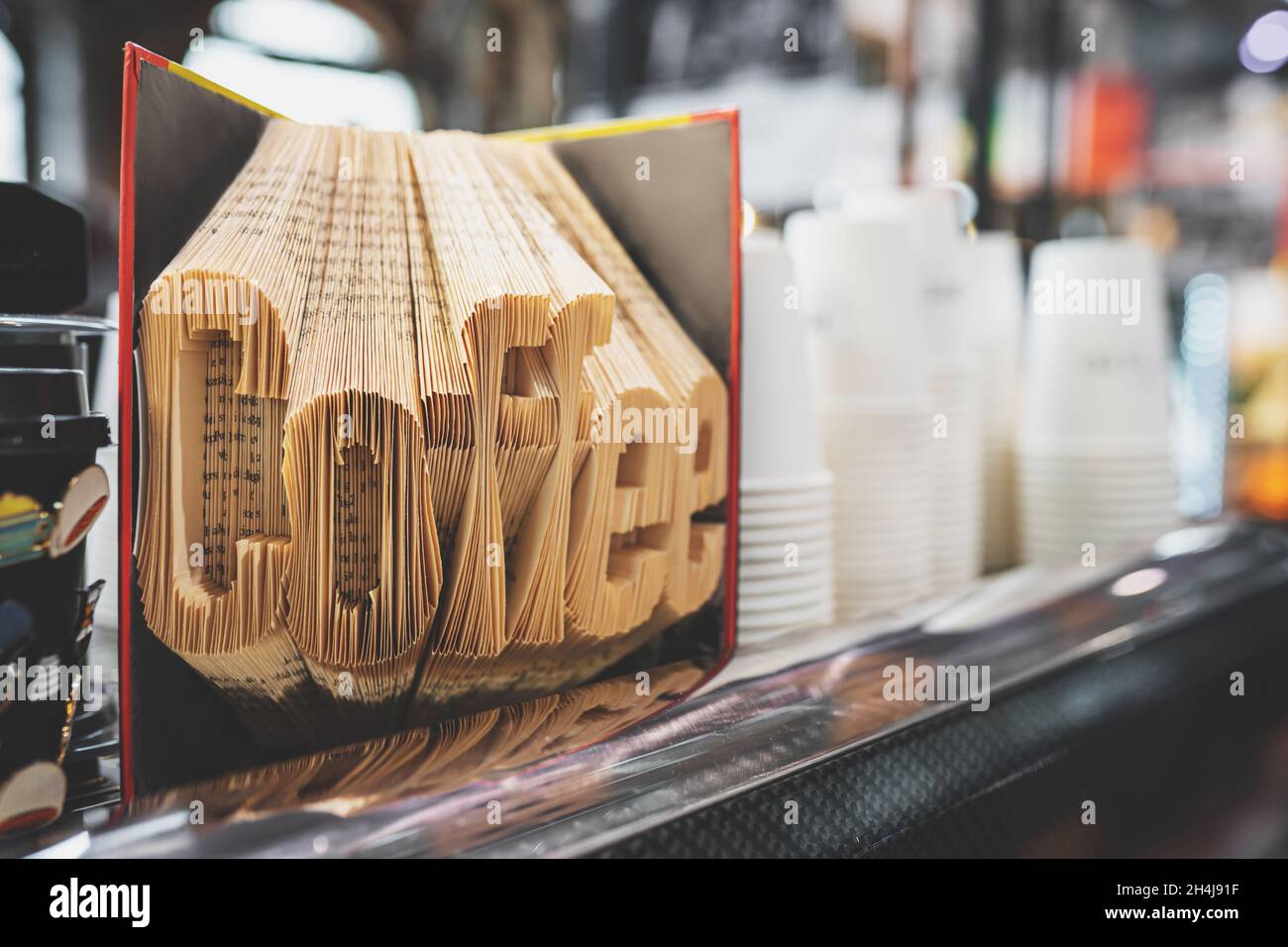 Word Coffee with folded book pages. Book sculpture decoration in a cozy coffee shop interior. White paper cups on the background. Toned image with selective focus. Coffee takeaway business concept Stock Photo