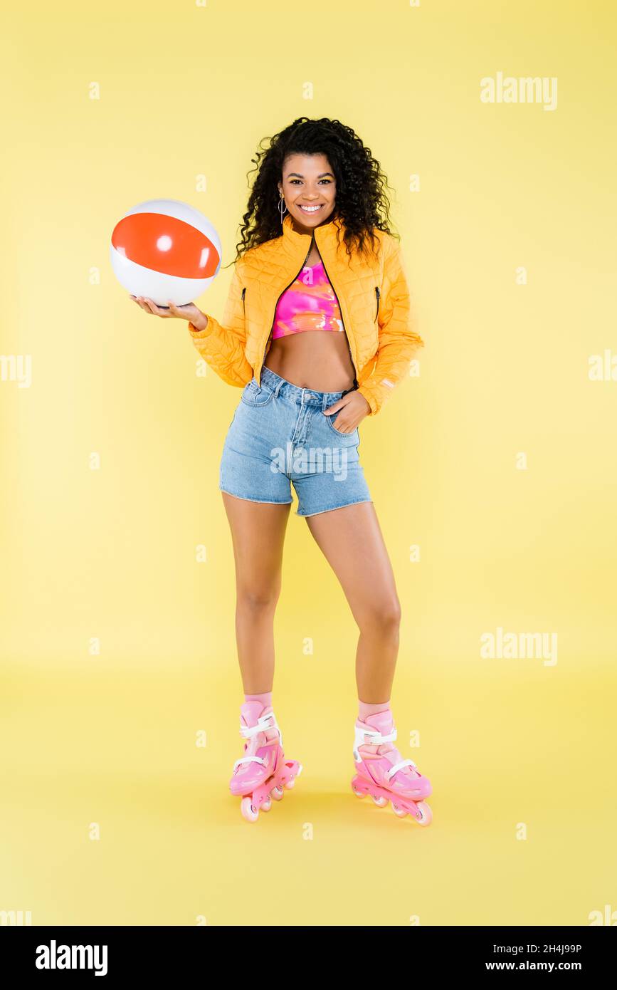 full length of positive african american young woman on roller skates holding inflatable ball on yellow Stock Photo