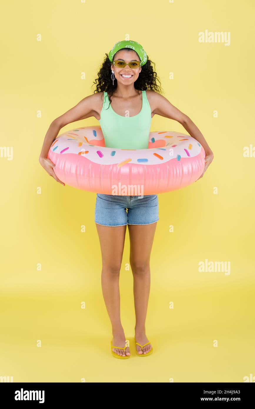 full length of joyful african american young woman standing with inflatable ring on yellow Stock Photo