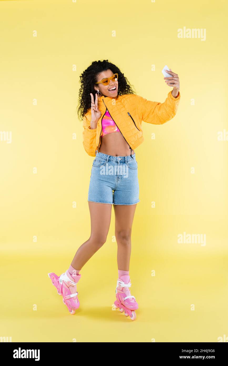 full length of amazed african american young woman on pink roller skates showing piece sign while taking selfie on yellow Stock Photo