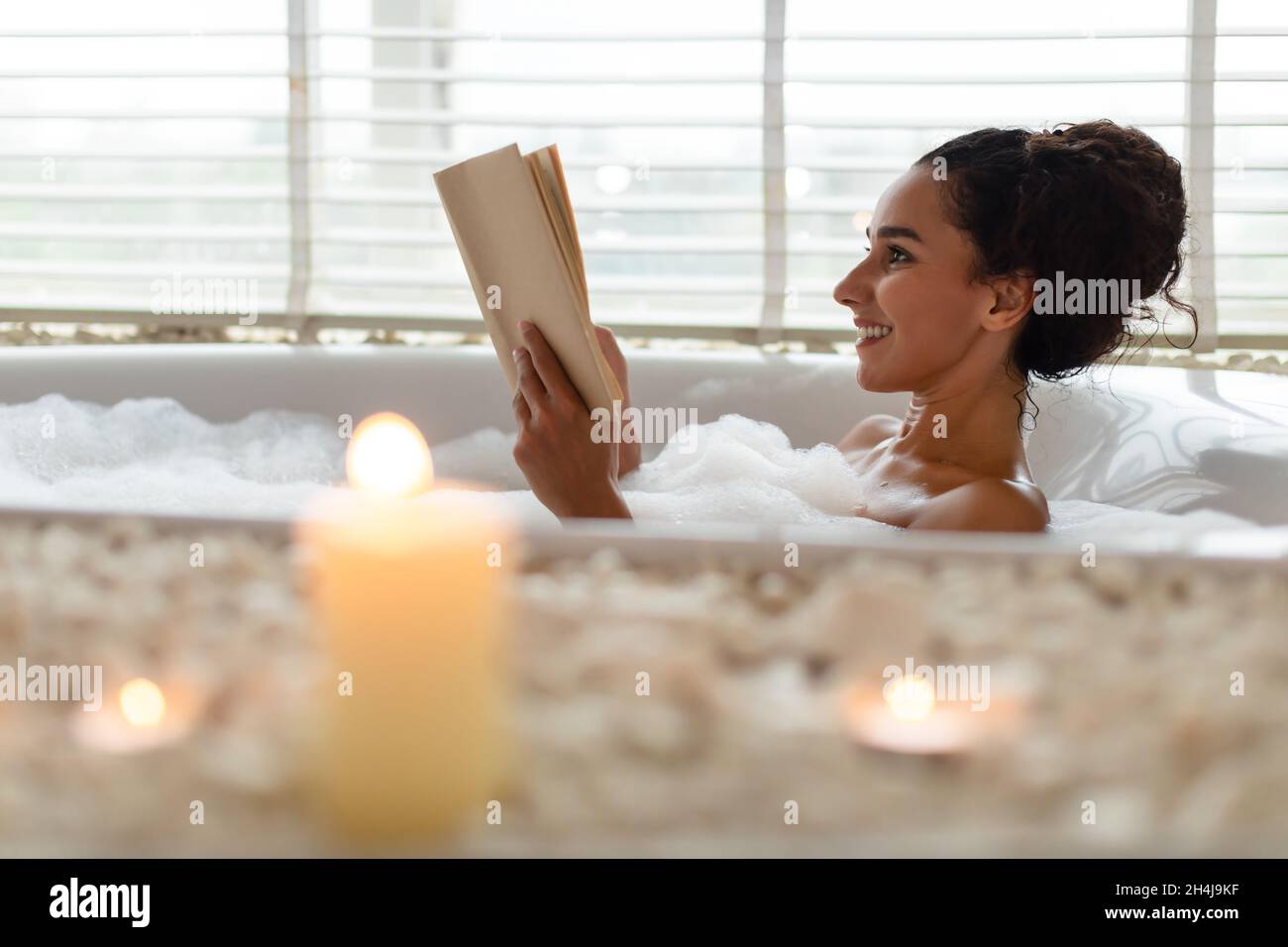 Lovely young woman lying in foamy bath, reading book in relaxing atmosphere with candles, indoors. Copy space Stock Photo
