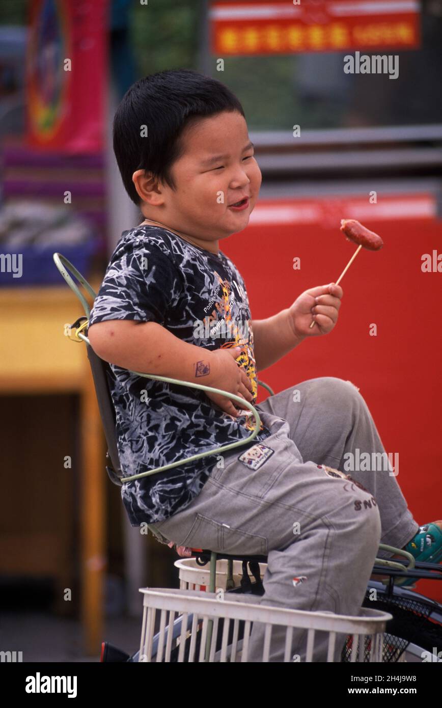 China 2000s. The Chinese one child policies operate in China unless there are exceptional circumstances. This over weight fat and indulged 'Little Emperor' happily eats a sausage on a stick while riding on the back of his mothers bicycle. 2001 HOMER SYKES Stock Photo