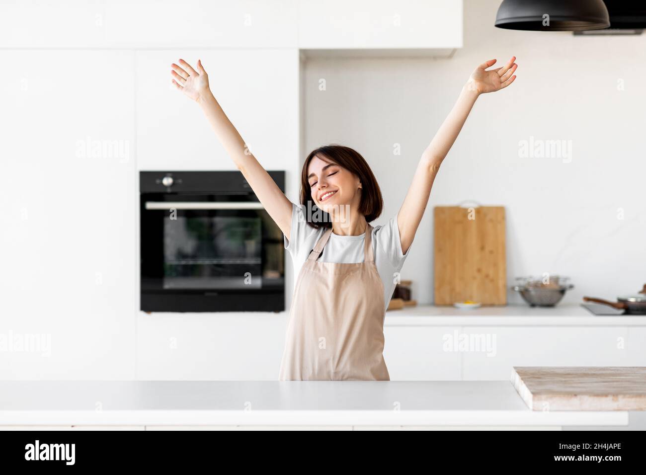 Relaxed young lady raising hands and feeling excited cooking alone in modern kitchen interior, having fun Stock Photo