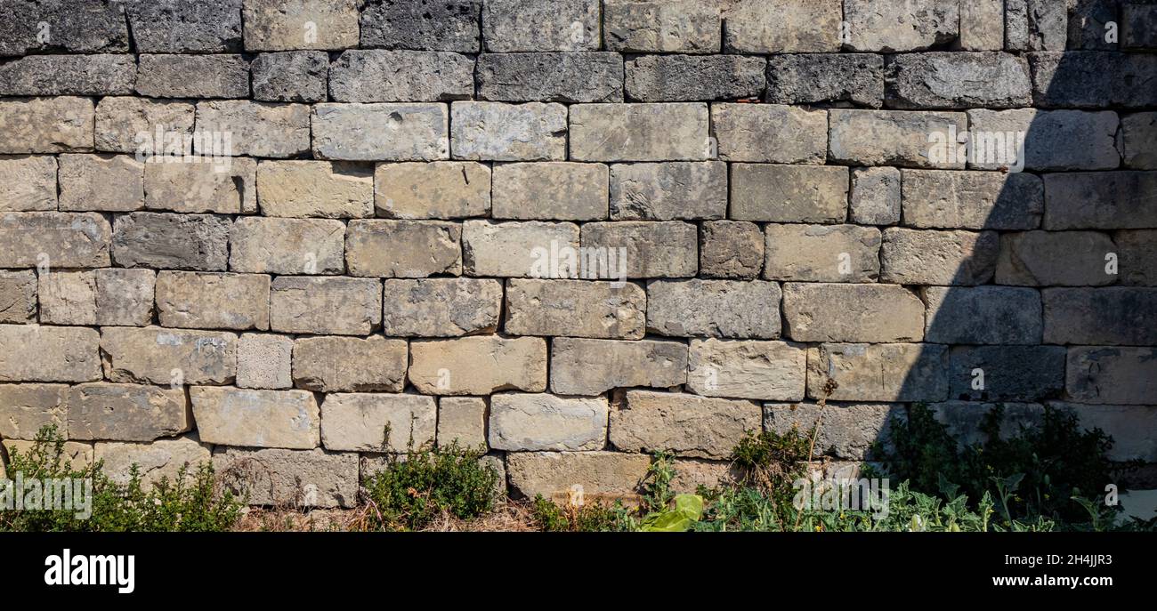 Texture, background, pattern. Ancient dry built stone brick wall. Irregular stones broken and ruined by time. Weeds grow low. Rough and dirty surface. Stock Photo