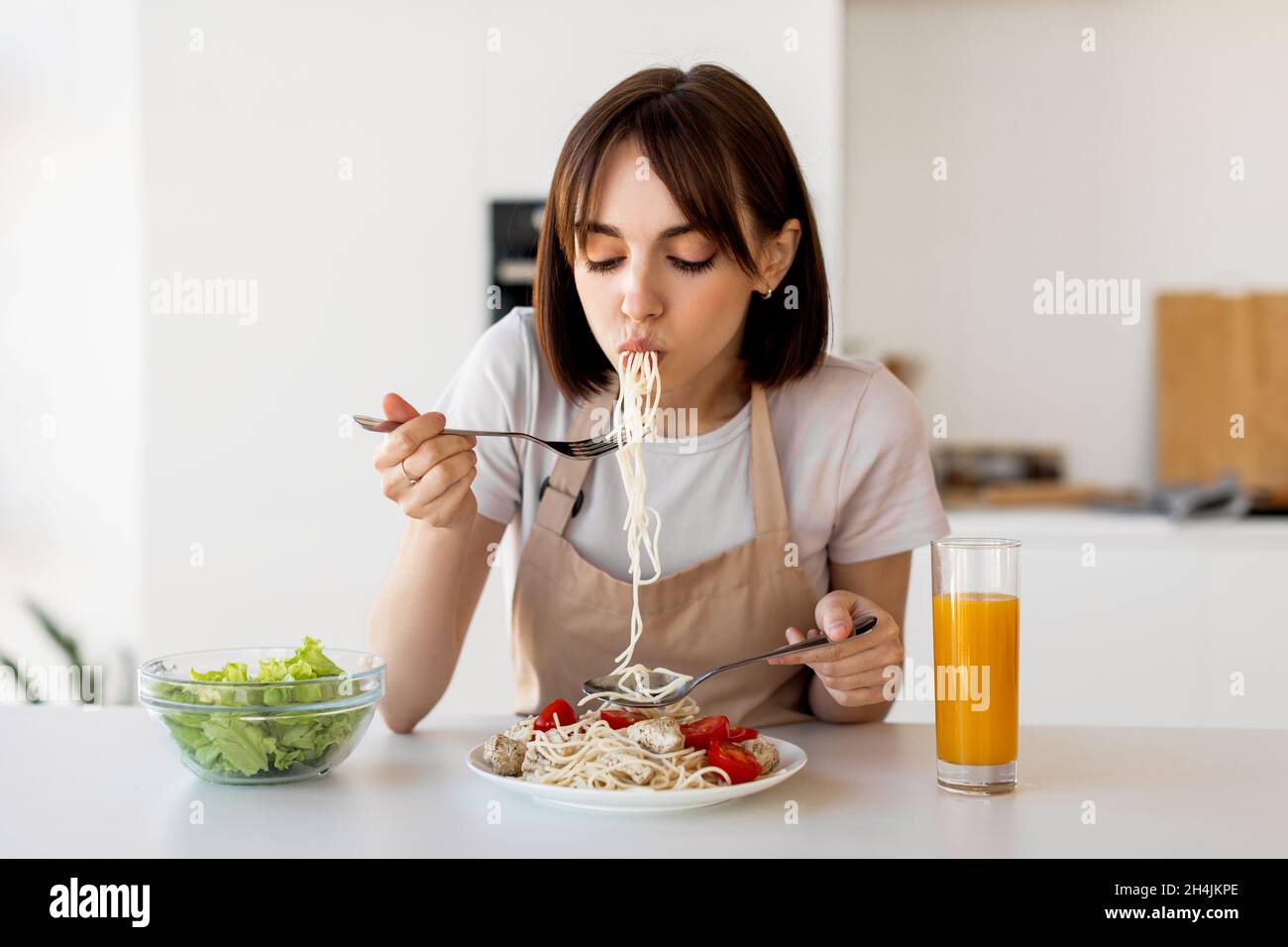 Young housewife tasting spaghetti and vegetable salad, enjoying delicious lunch at home, sitting in modern kitchen Stock Photo