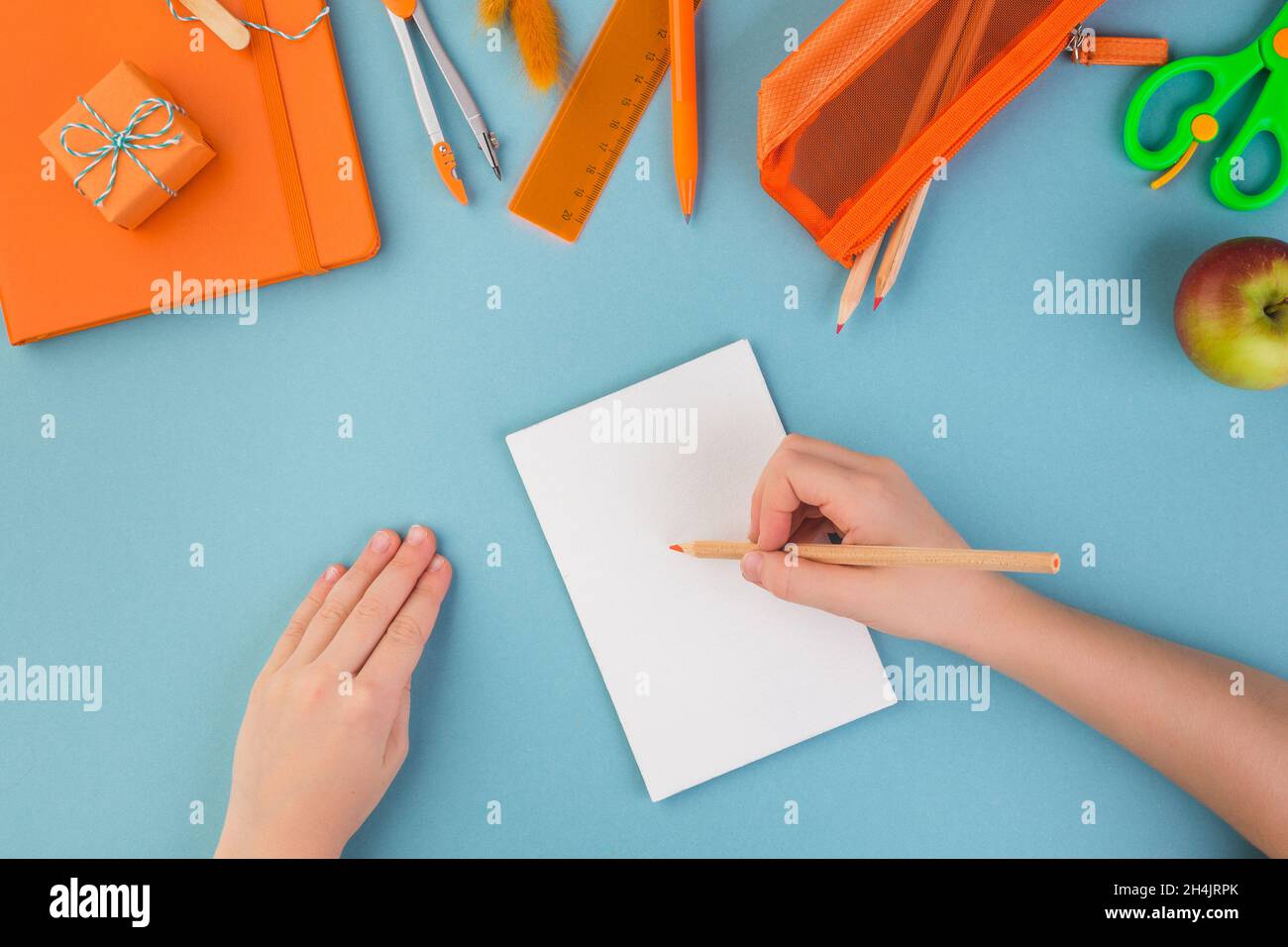 Child hand writes on white peace of paper school or office workspace with orange supplies on blue background. Flat lay. Stock Photo