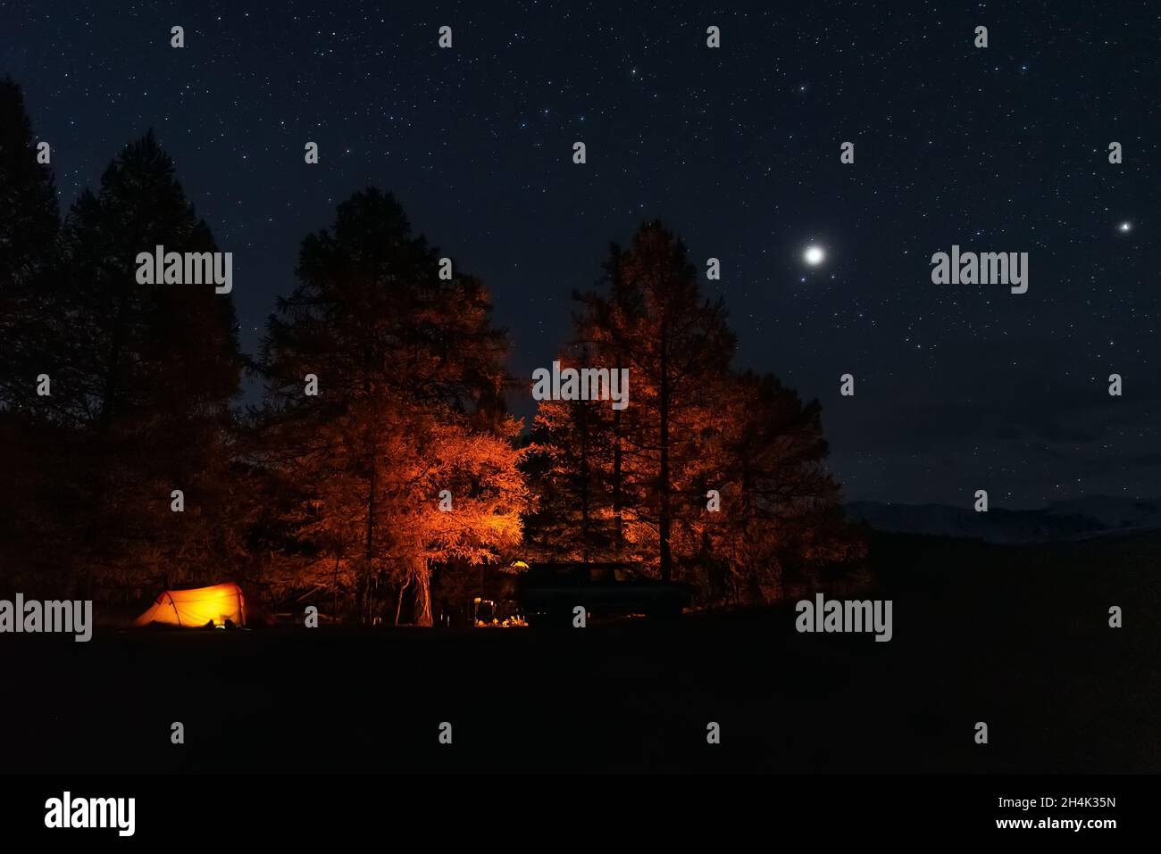 Scenic night view of tourist camp with illuminated orange tent, yellow larch trees, bonfire and car on top in mountains against starry sky Stock Photo