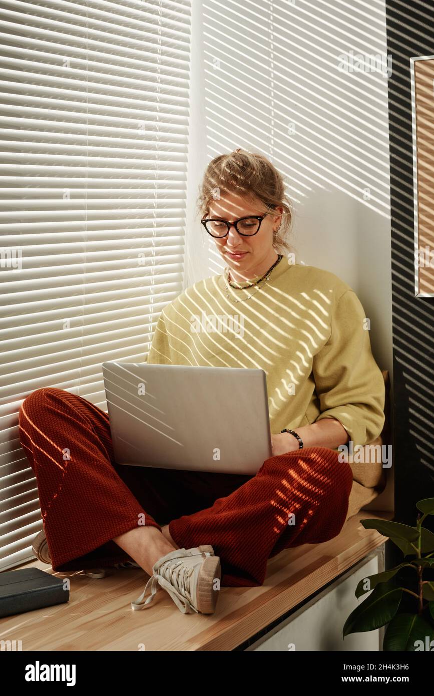 Young woman in eyeglasses sitting on windowsill and communicating online using her laptop Stock Photo