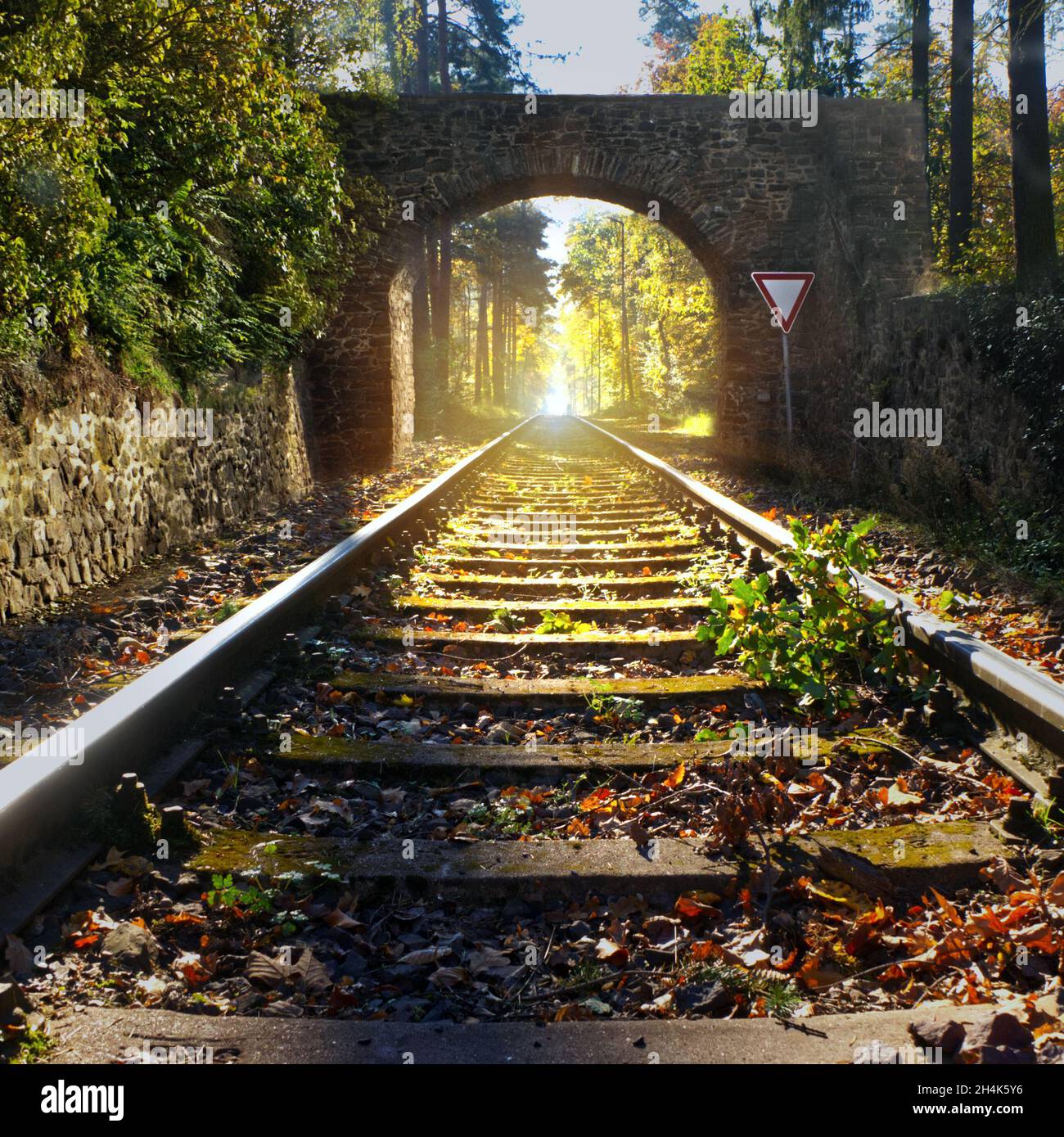 Rails lead in central perspective through an old arched gate made of natural stone to the glistening sun in the background, composite image Stock Photo