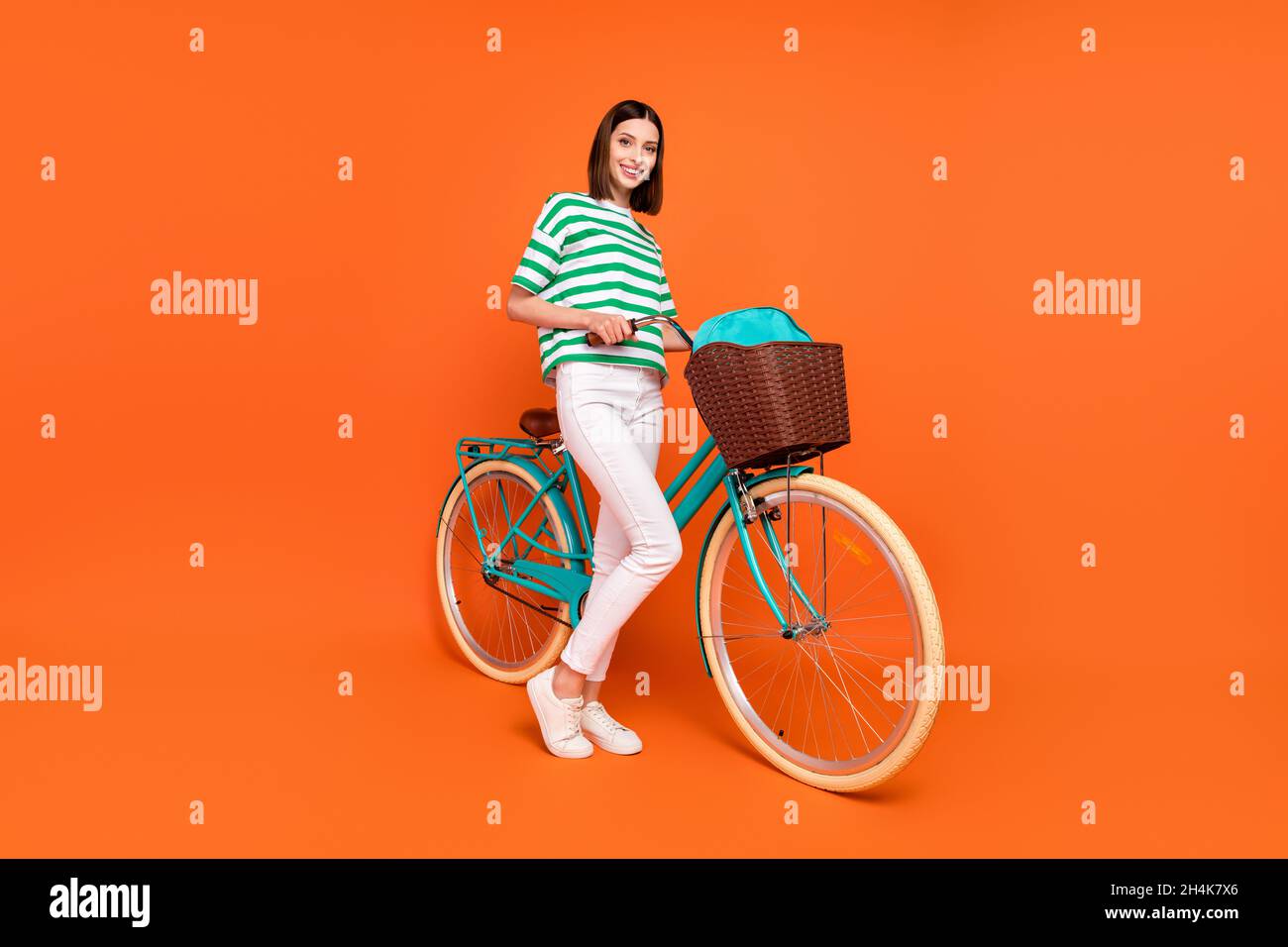 Full size photo of cute brunette young lady travel on bicycle wear t-shirt trousers sneakers isolated on orange color background Stock Photo