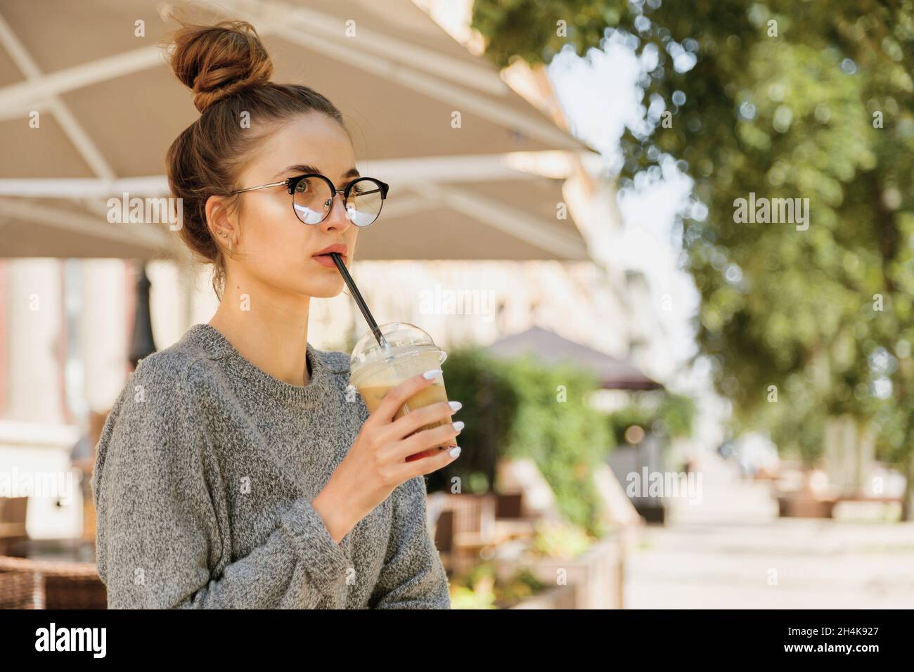 Alone young woman in eyeglasses drinking milkshake and looking away in cozy cafe outdoor. Stock Photo