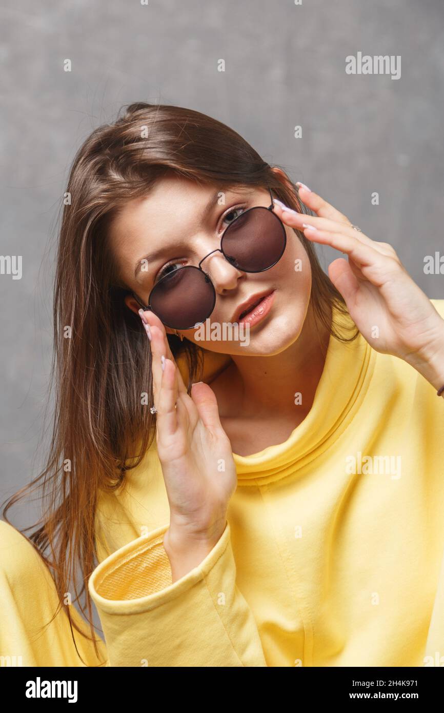 Young woman in yellow sweatshirt and sunglasses on a gray background. Stock Photo