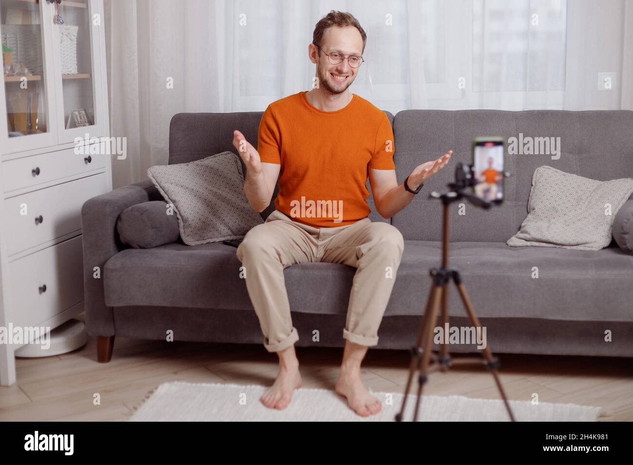 Hipster man blogger recording vlog on mobile phone sit on couch in living room and explain and gesturing with hands. Stock Photo