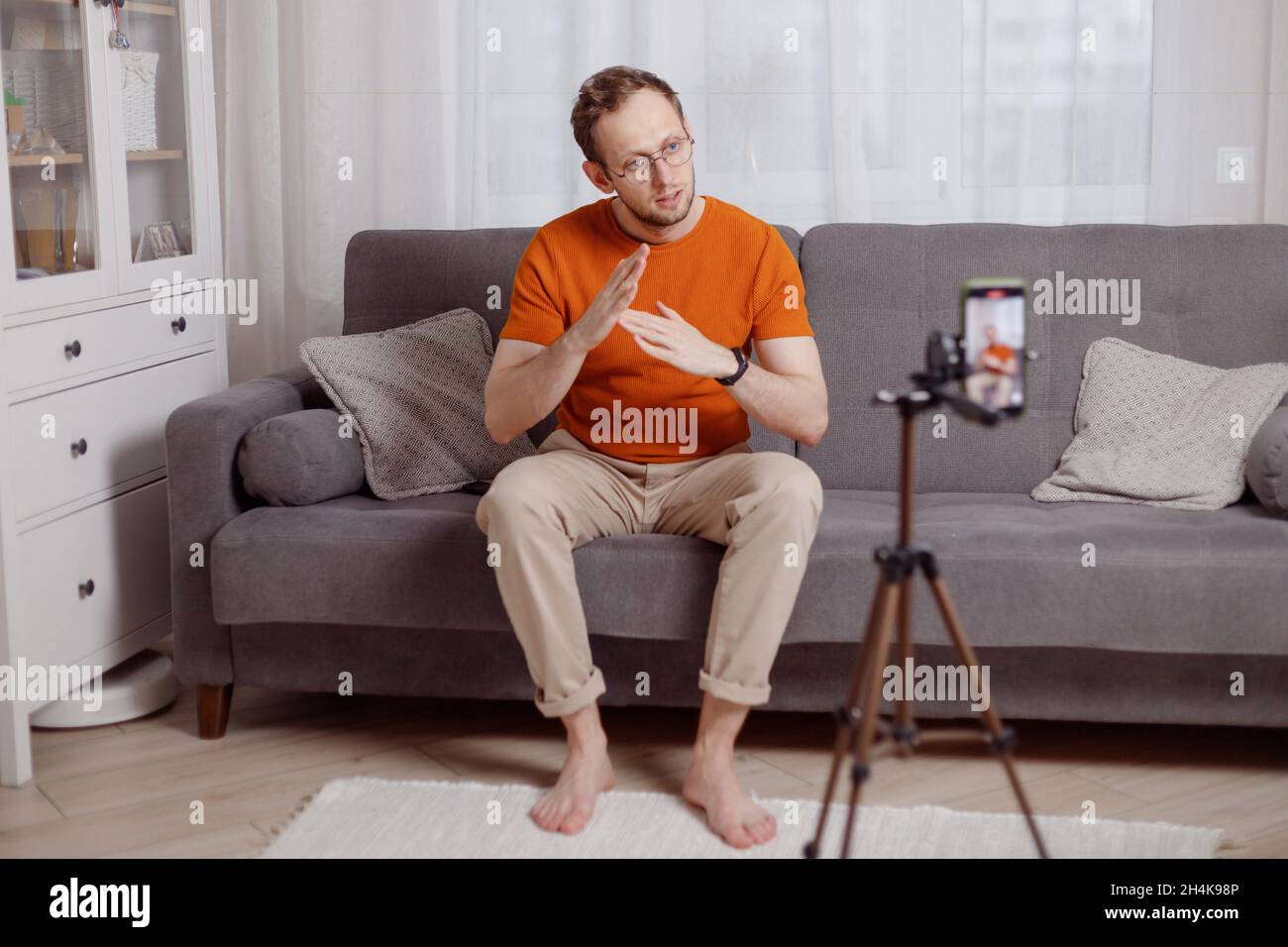 Hipster man blogger recording vlog on mobile phone sit on couch in living room and explain and gesturing with hands. Stock Photo