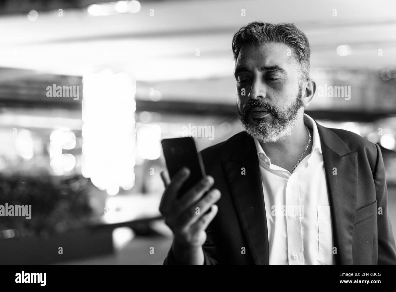 Black and white portrait of handsome Indian businessman in city at night using mobile phone Stock Photo