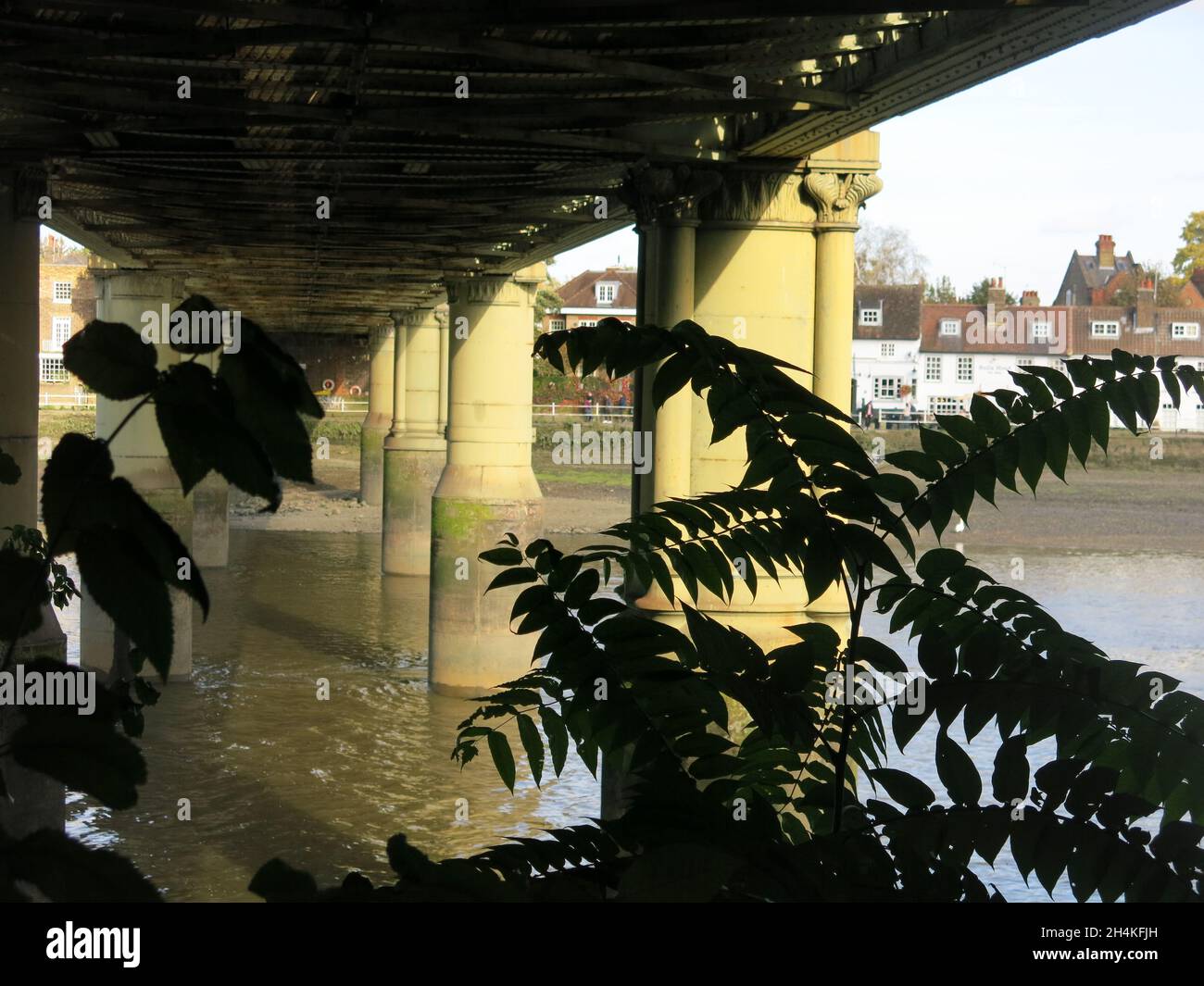 View looking underneath the line of the Kew Railway Bridge towards the north side of the River Thames with sunlight on support pillars in the water. Stock Photo