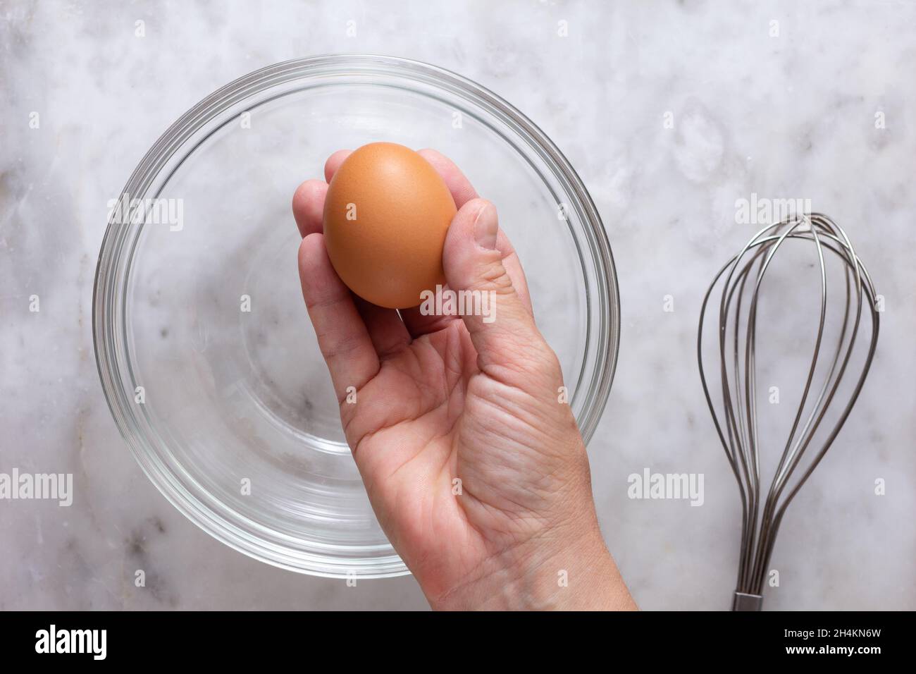 Top view of right woman hand holding one raw brown egg above glass bowl and whisk on the marble surface Stock Photo