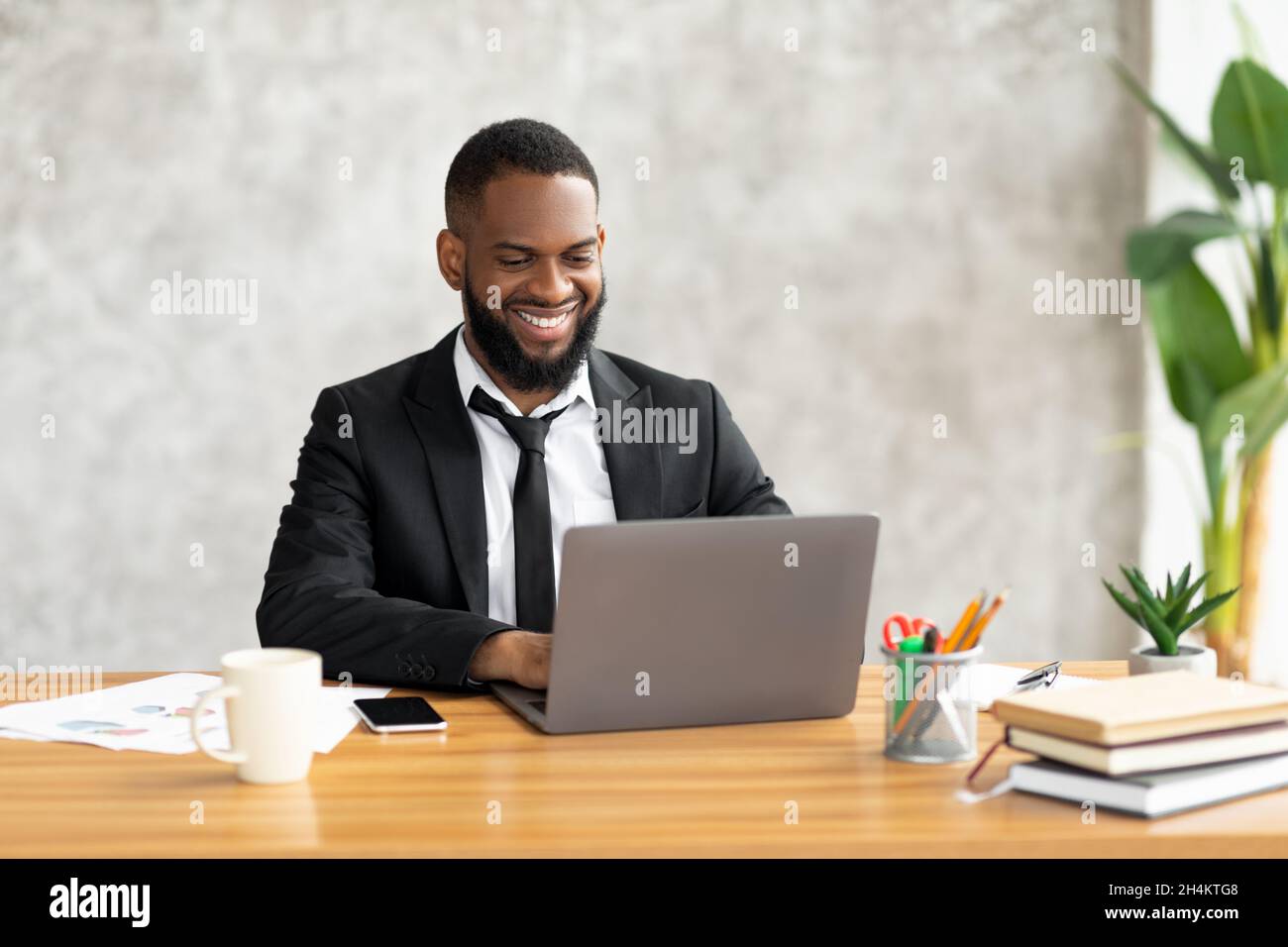 Portrait of handsome smiling African American male entrepreneur in suit working on laptop at desk in modern office, using computer at workplace, typin Stock Photo