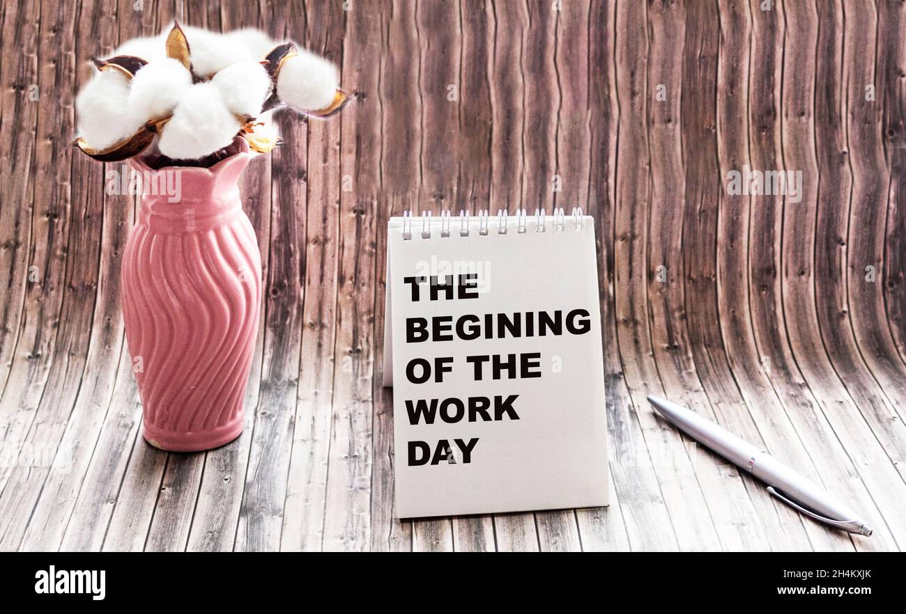 the text THE BEGINNING OF THE WORK DAY is written on a notebook, next to a bouquet of flowers on a wooden background. Stock Photo