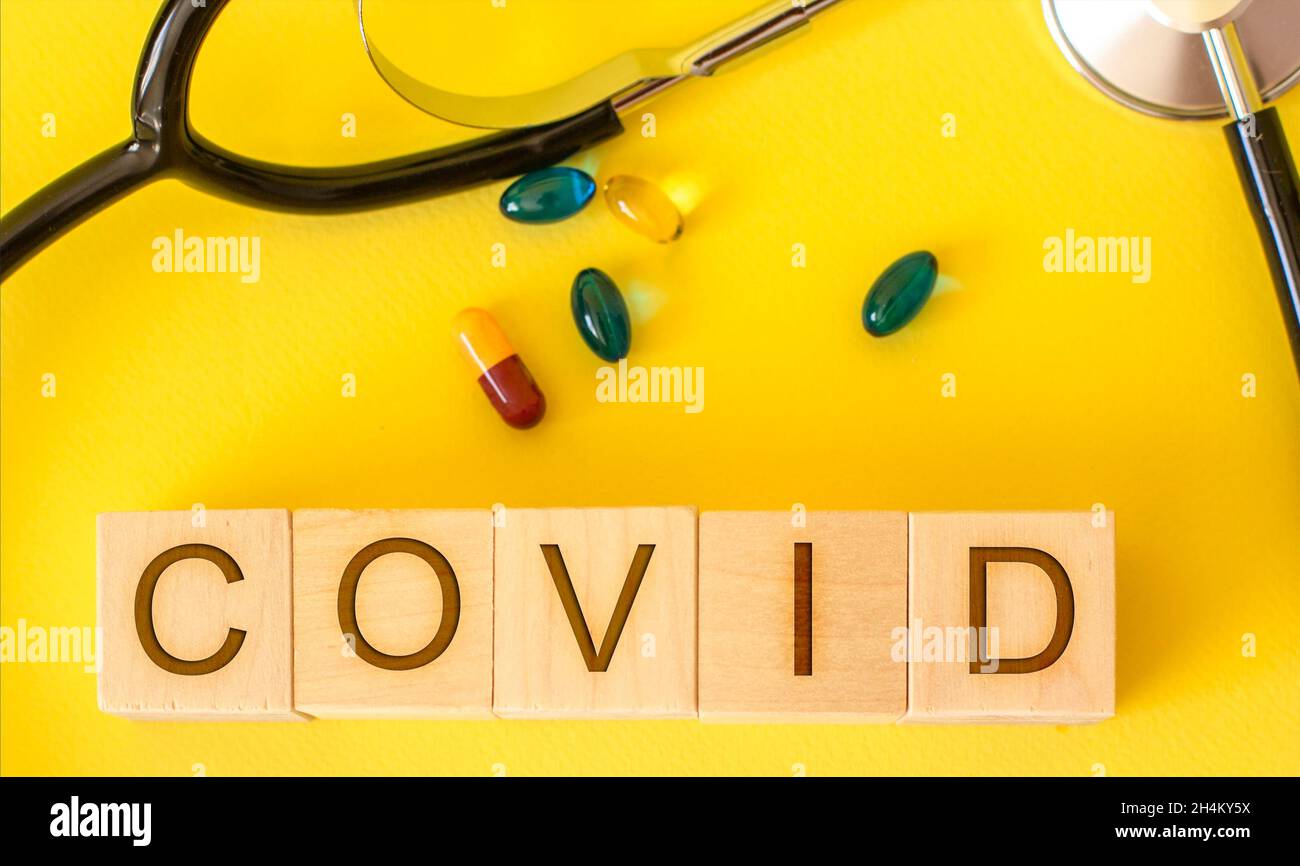 Wooden cubes with the word COVID on a yellow background with pills and with a stethoscope Stock Photo