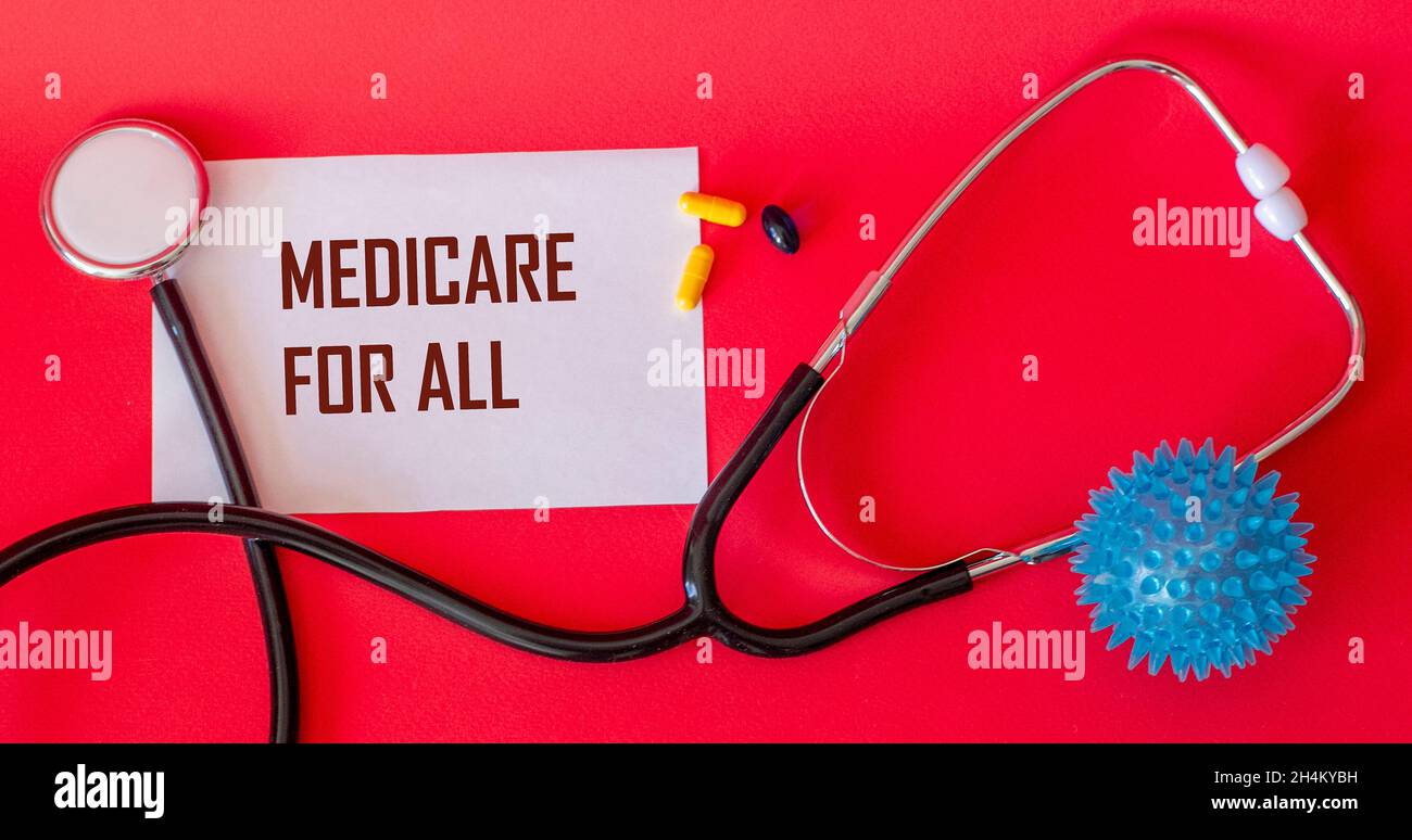 MEDICINE FOR ALL, the text is written on a notebook. A stethoscope, pills and a dummy virus lie on a red background Stock Photo