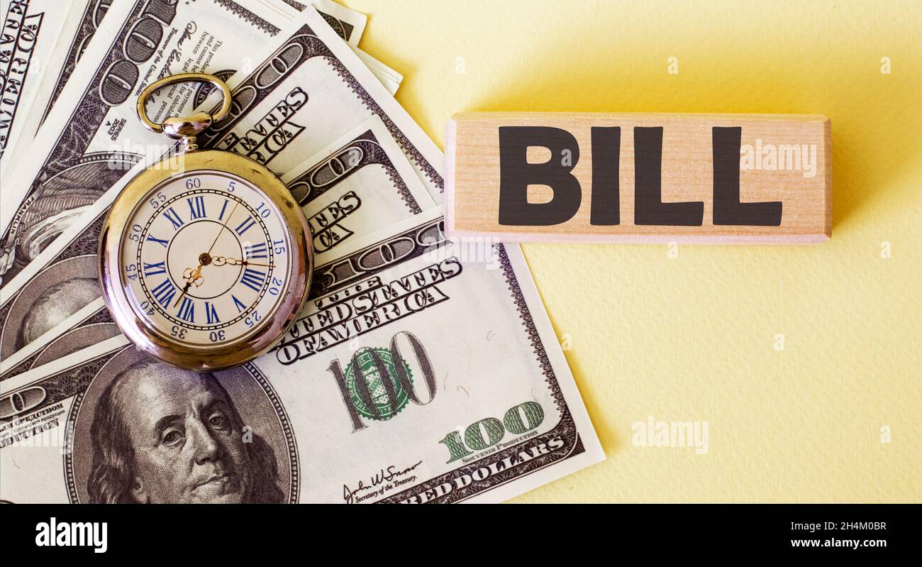 The word Bill is written on a wooden block, a stack of 100 dollars and a clock on a yellow background. American currency. Money and financial concept. Stock Photo