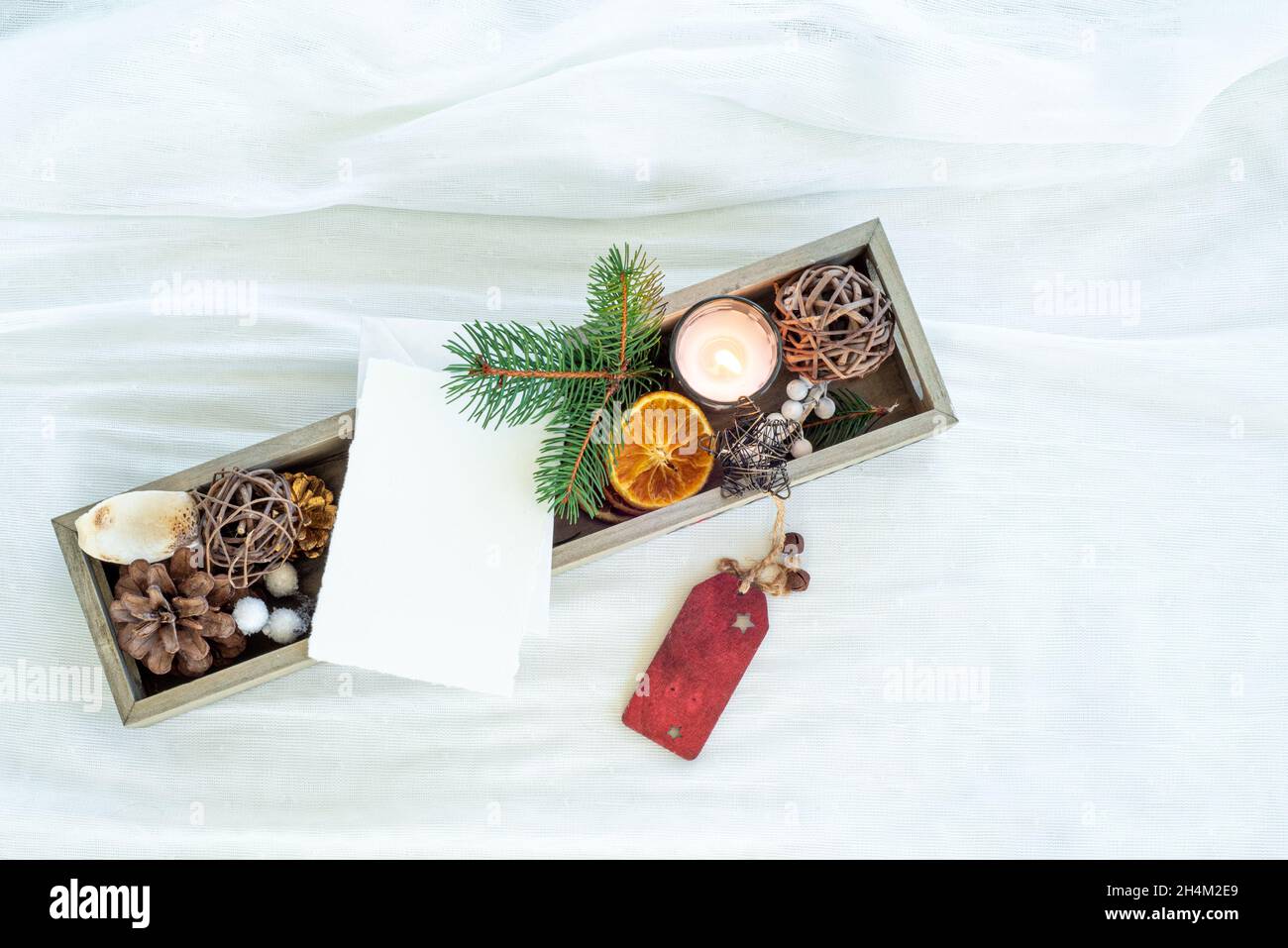 Christmas decorations in small wooden box. Blank greeting card mockup among fir twig, candle, dried citrus slice, pine cone, and ornamental balls Stock Photo