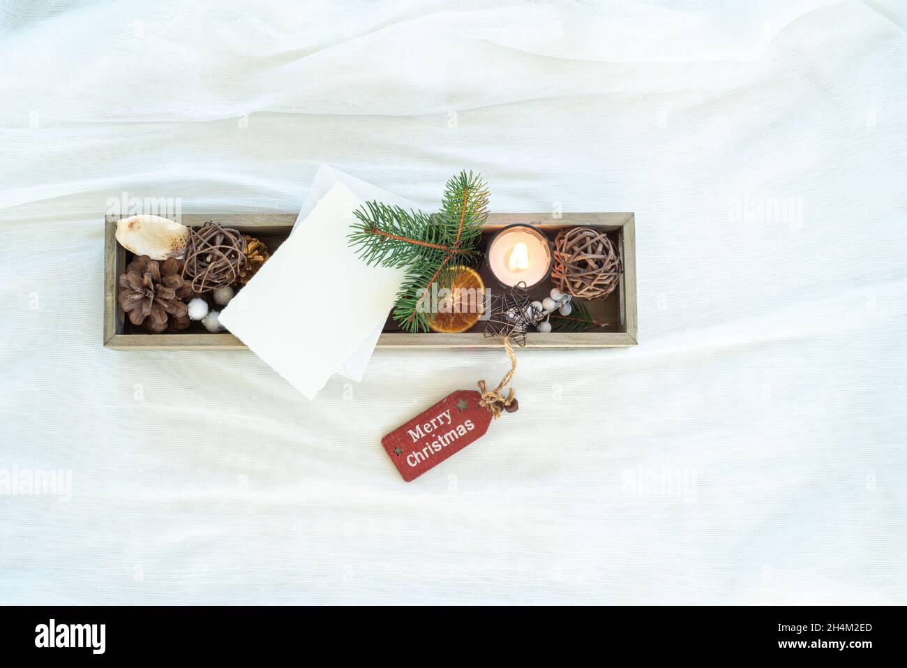 Christmas decorations in small wooden box. Blank greeting card mockup among fir twig, candle, dried citrus slice, pine cone, and ornamental balls Stock Photo