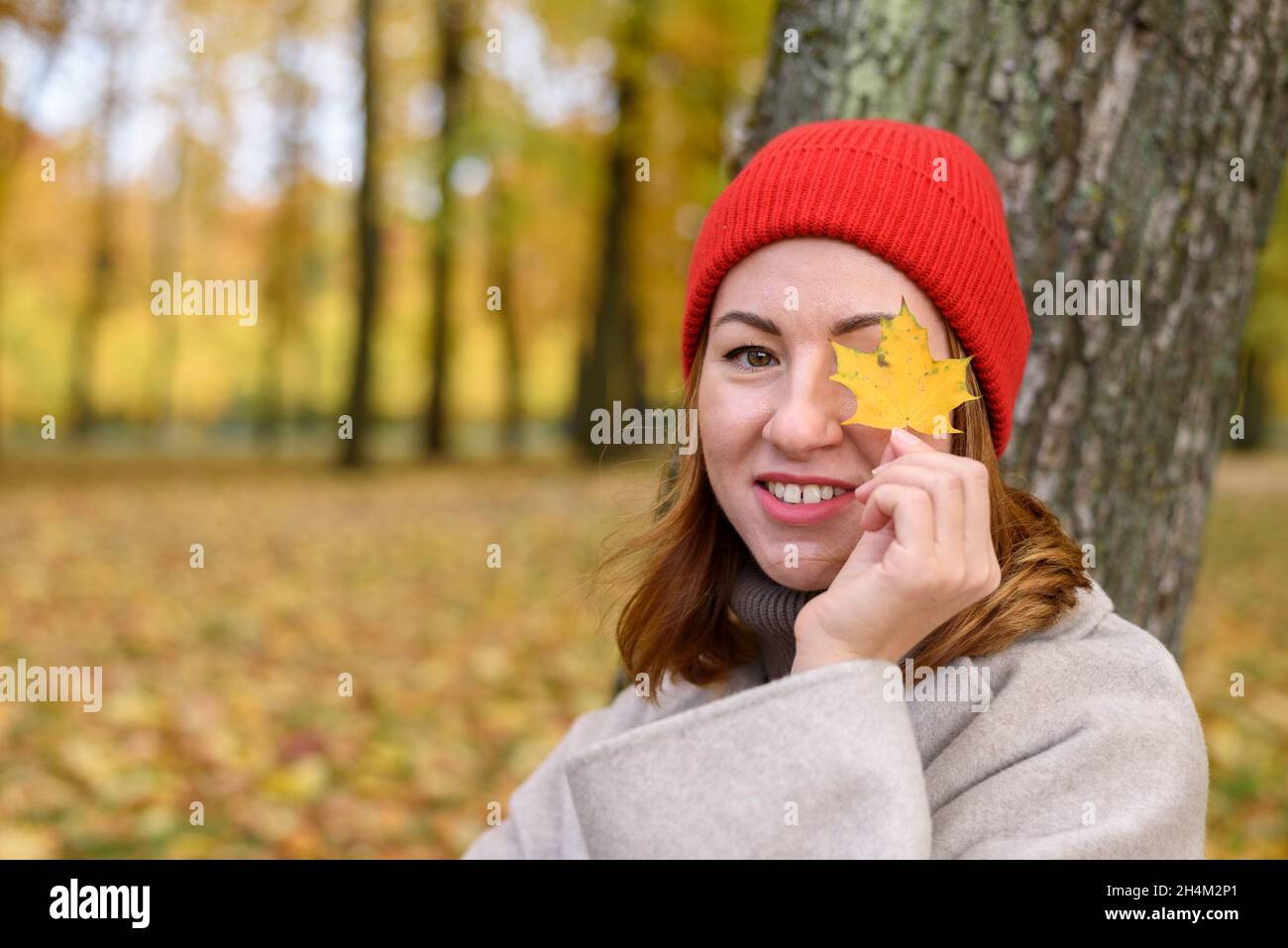 Woman in red knitted hat holding maple leaf near her eye at autumn park Stock Photo