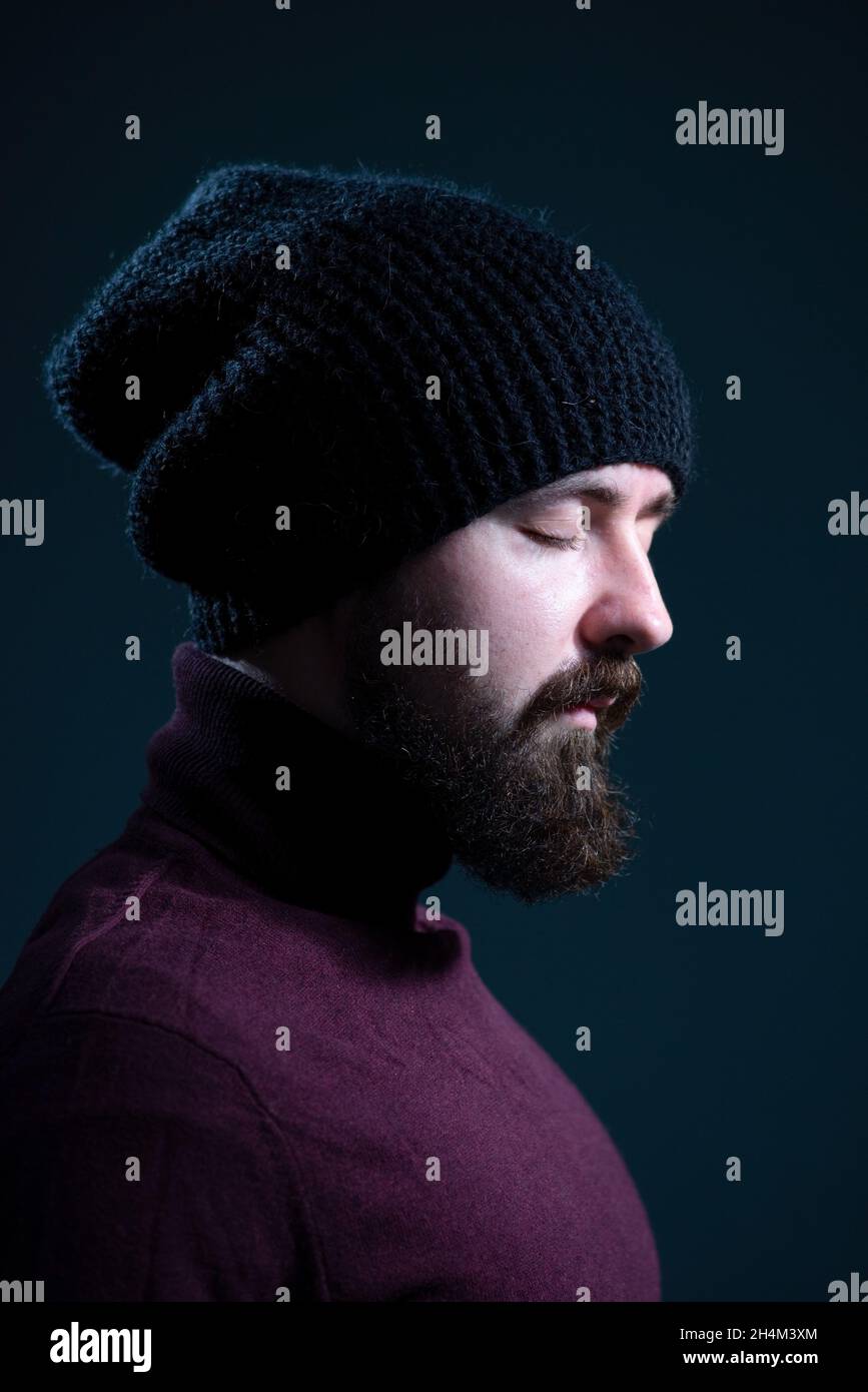 bearded guy with closed eyes in a knitted black hat and burgundy sweater. On a gray background. Stock Photo