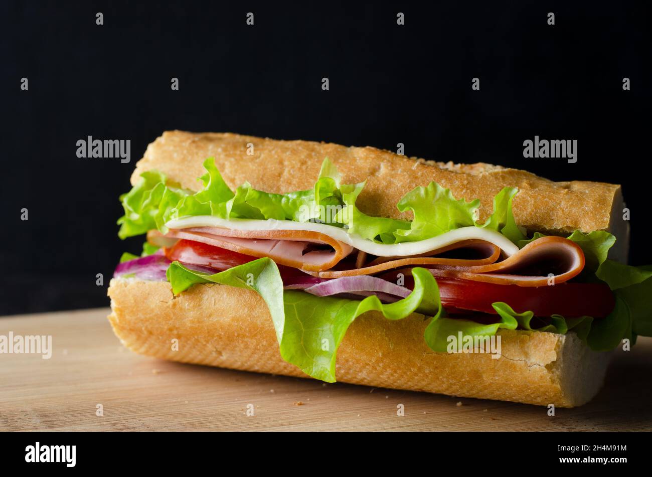 Fresh submarine sandwich with ham, cheese, bacon, tomatoes, lettuce and onions on dark wooden background Stock Photo