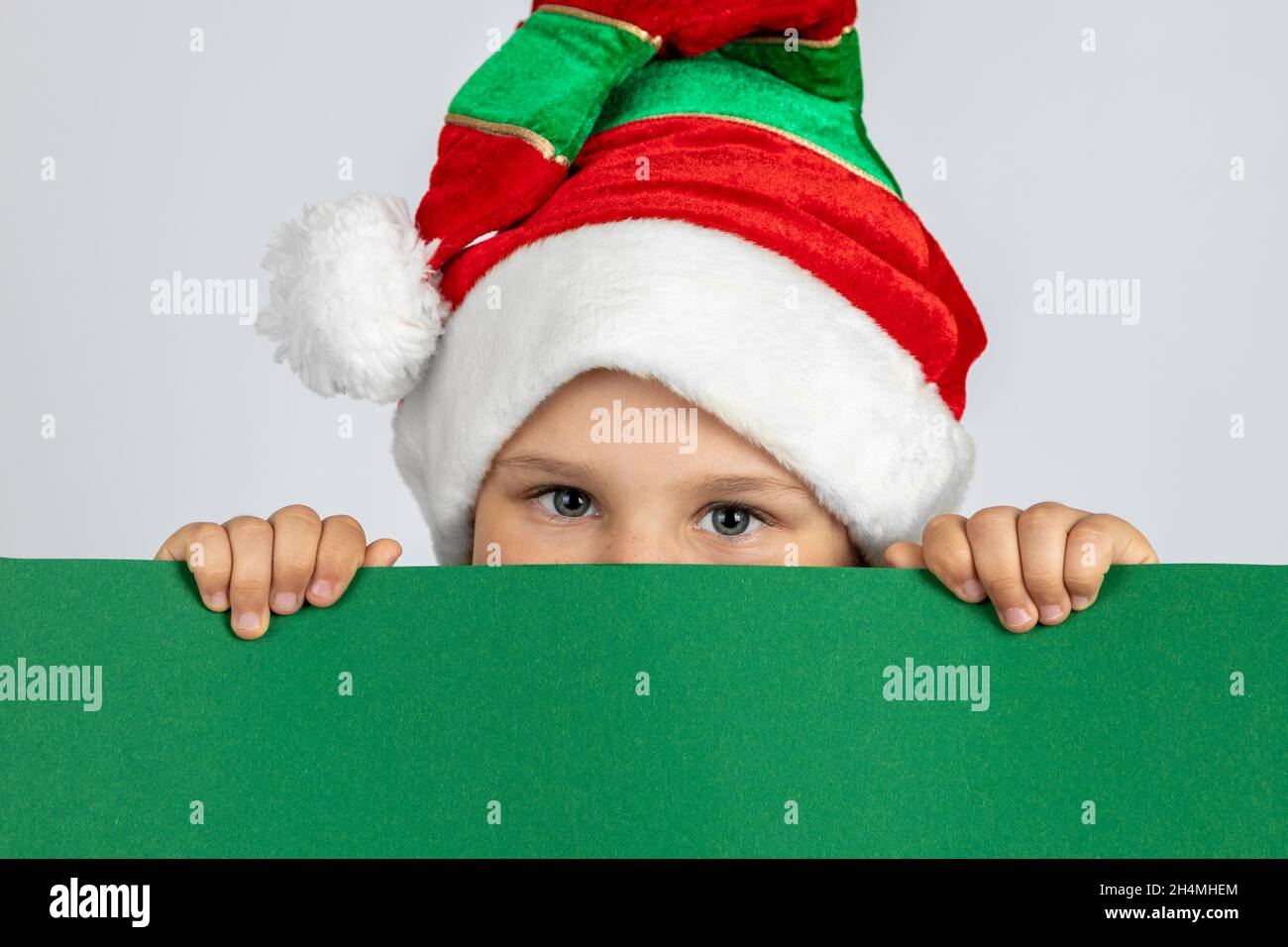 close-up portrait of mischievous child in Christmas gnome hat hiding behind black green poster with copy space in hands, isolated on white background Stock Photo