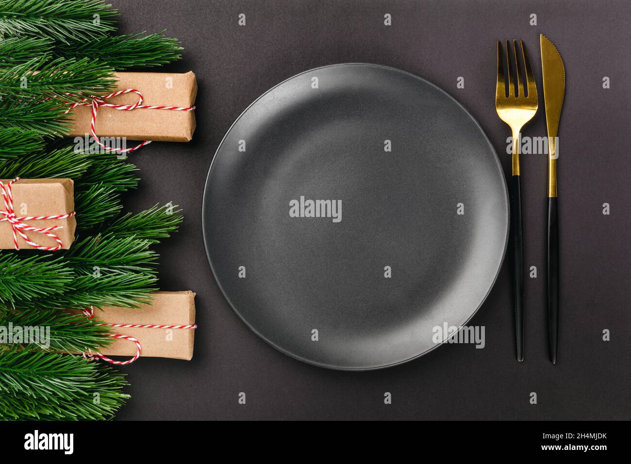 top view of black cutlery knife and fork near black empty plate on black table with Christmas tree branches and decorations from gift boxes Stock Photo